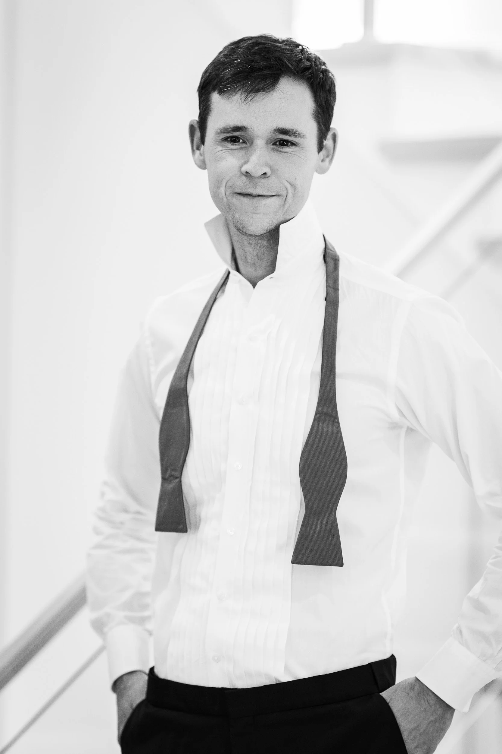Black and white photo of a man wearing a white dress shirt and dark pants with a dark apron, standing in front of a staircase with a handrail. He has short, dark hair, and is smiling slightly with one hand in his pocket.