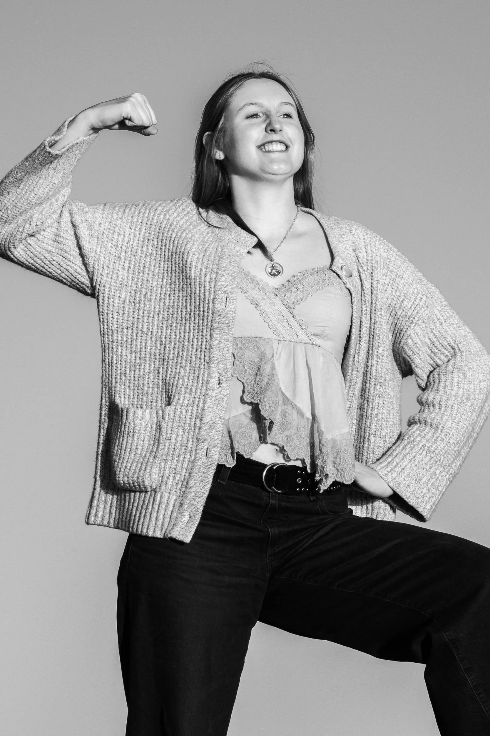 A young woman with long hair smiling and flexing her right arm, wearing a cardigan, lace top, and black pants, in a black-and-white studio photo.