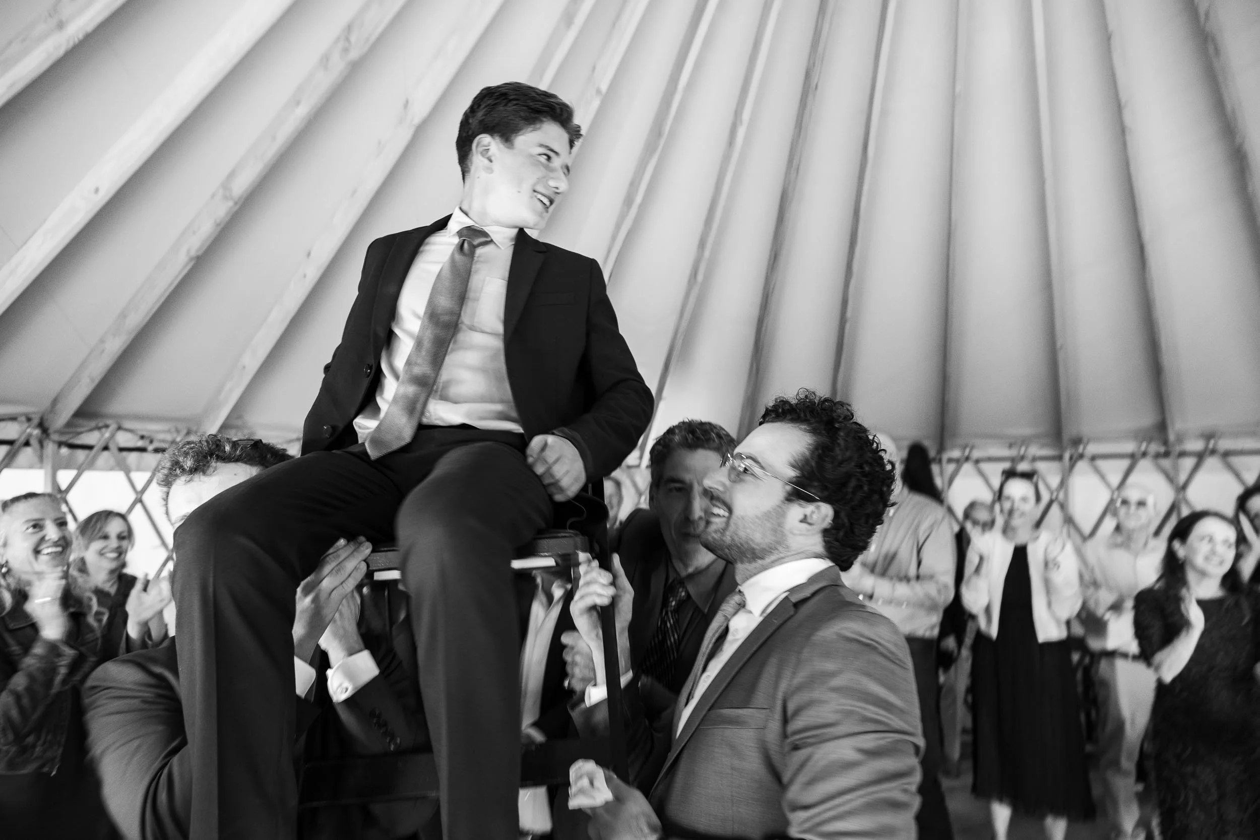 A man in a suit is sitting on a chair held up by several people. He appears to be smiling or laughing, and others around him, who are also dressed in formal attire, are smiling, clapping, or looking at him. The setting seems to be a celebratory event