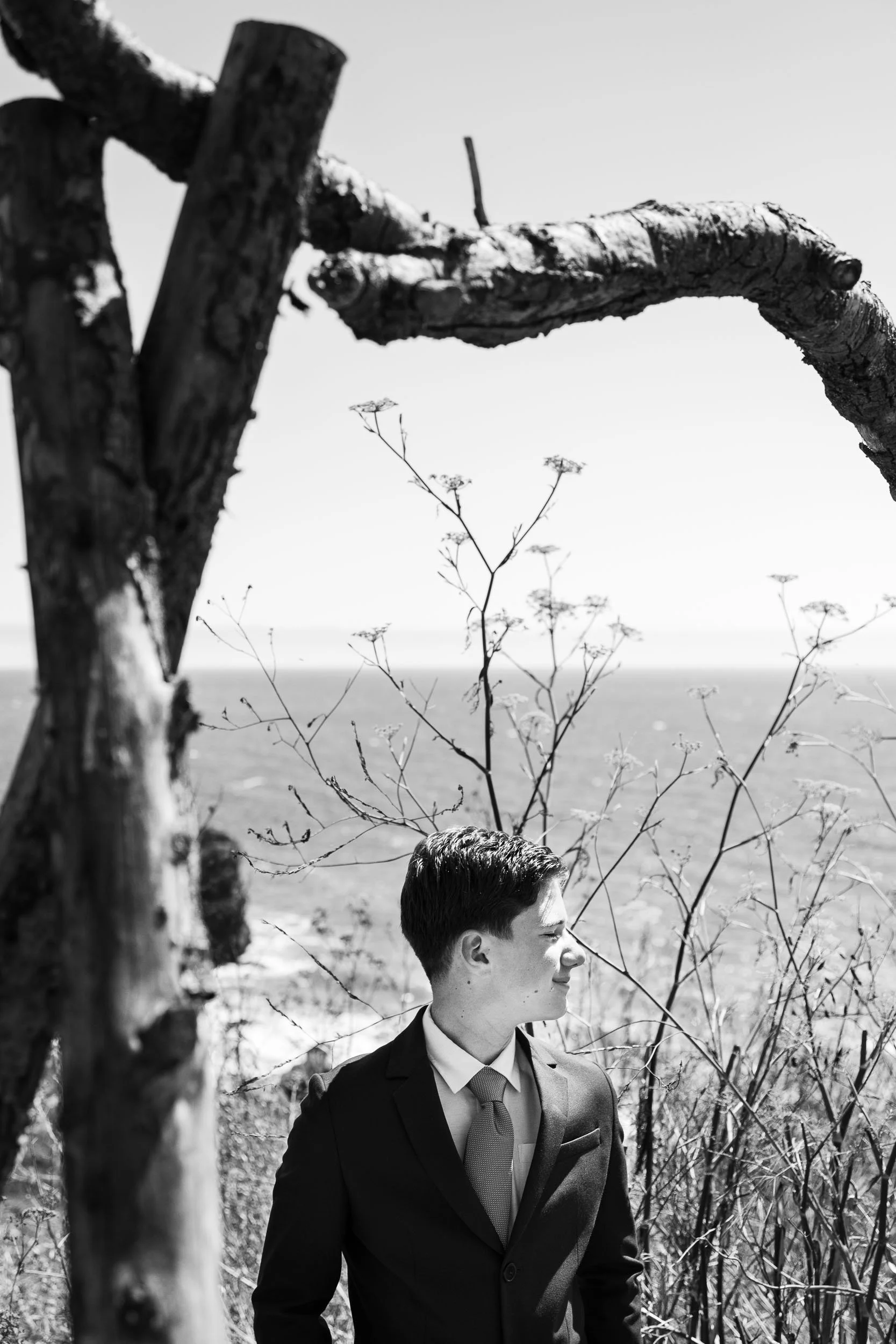 A young man in a suit looking to the right with eyes closed, standing outdoors among tall grasses and a barren tree, with the ocean in the background, in black and white.