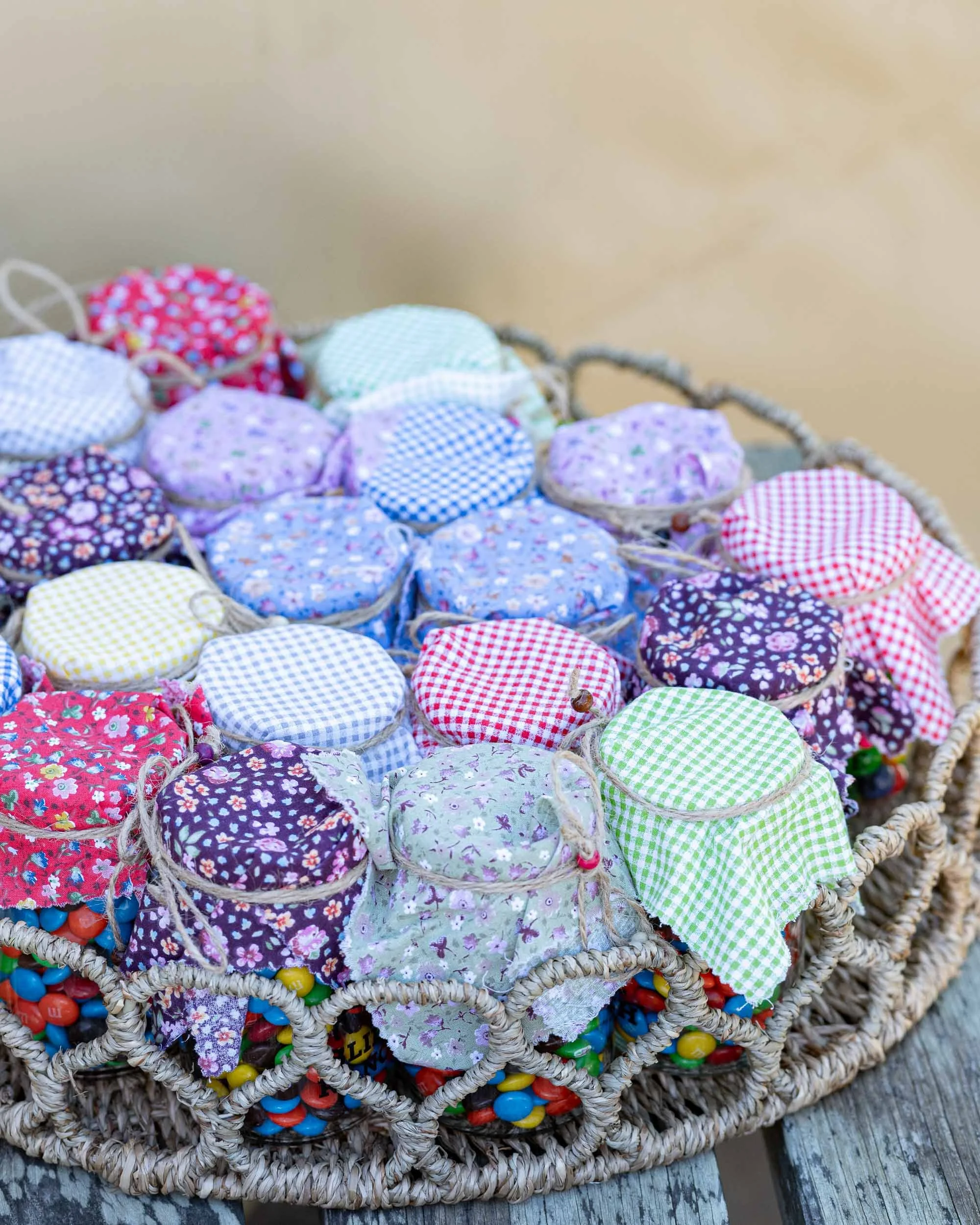 A wicker basket filled with small fabric-covered jars with drawstring lids, decorated in various colorful floral and checkered patterns, and filled with colorful candies.