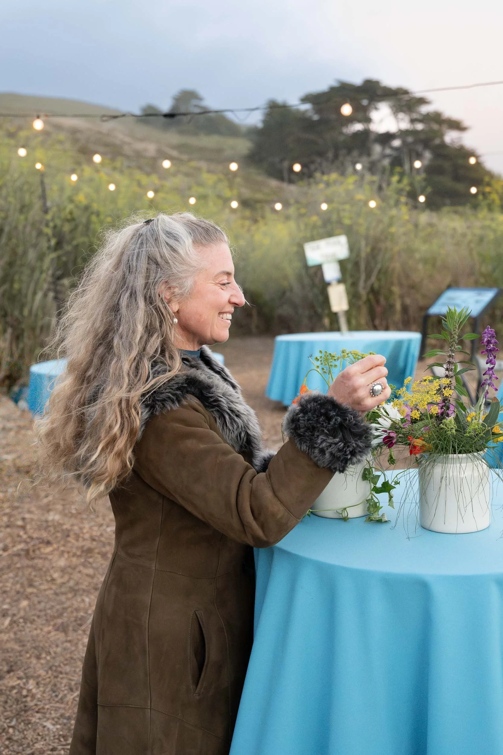 A woman with gray curly hair in a brown coat with fur accents, smiling and arranging flowers on a blue table outdoors at dusk, with string lights hanging above, hills and trees in the background.
