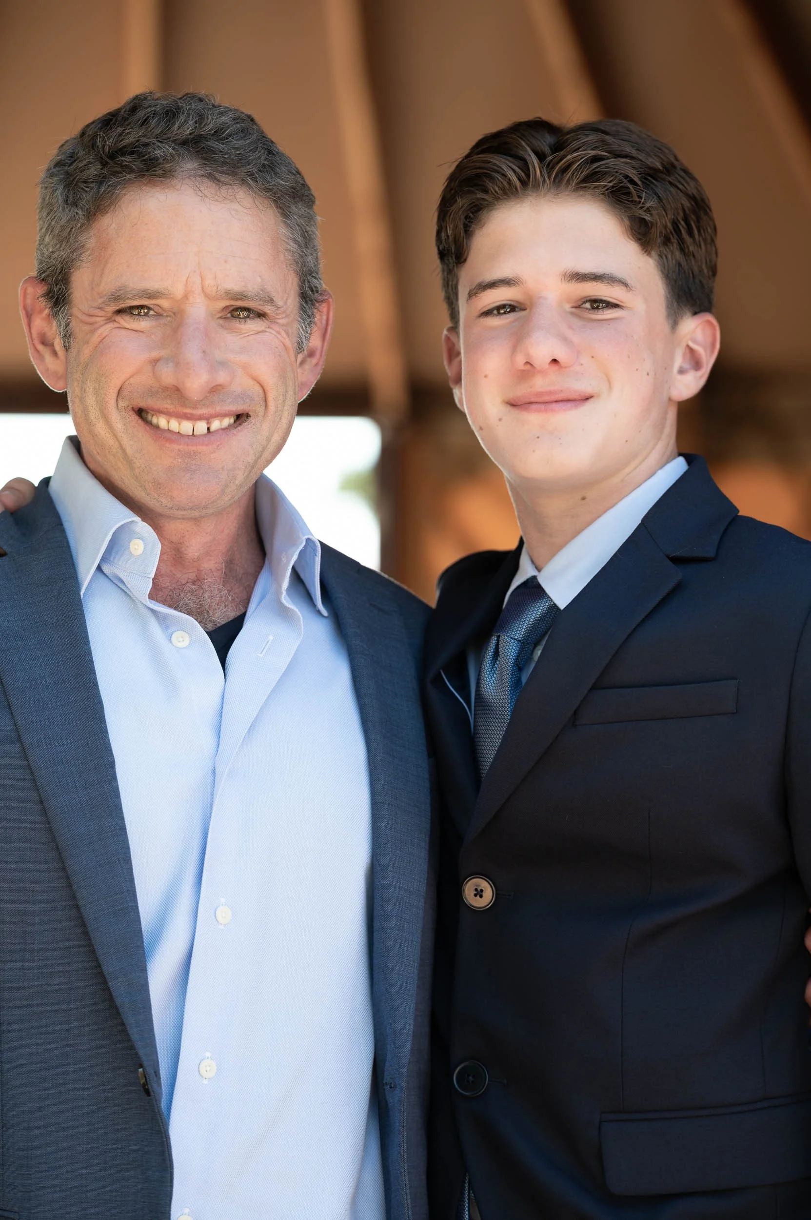 A smiling older man and a younger man in suits standing closely together outdoors.