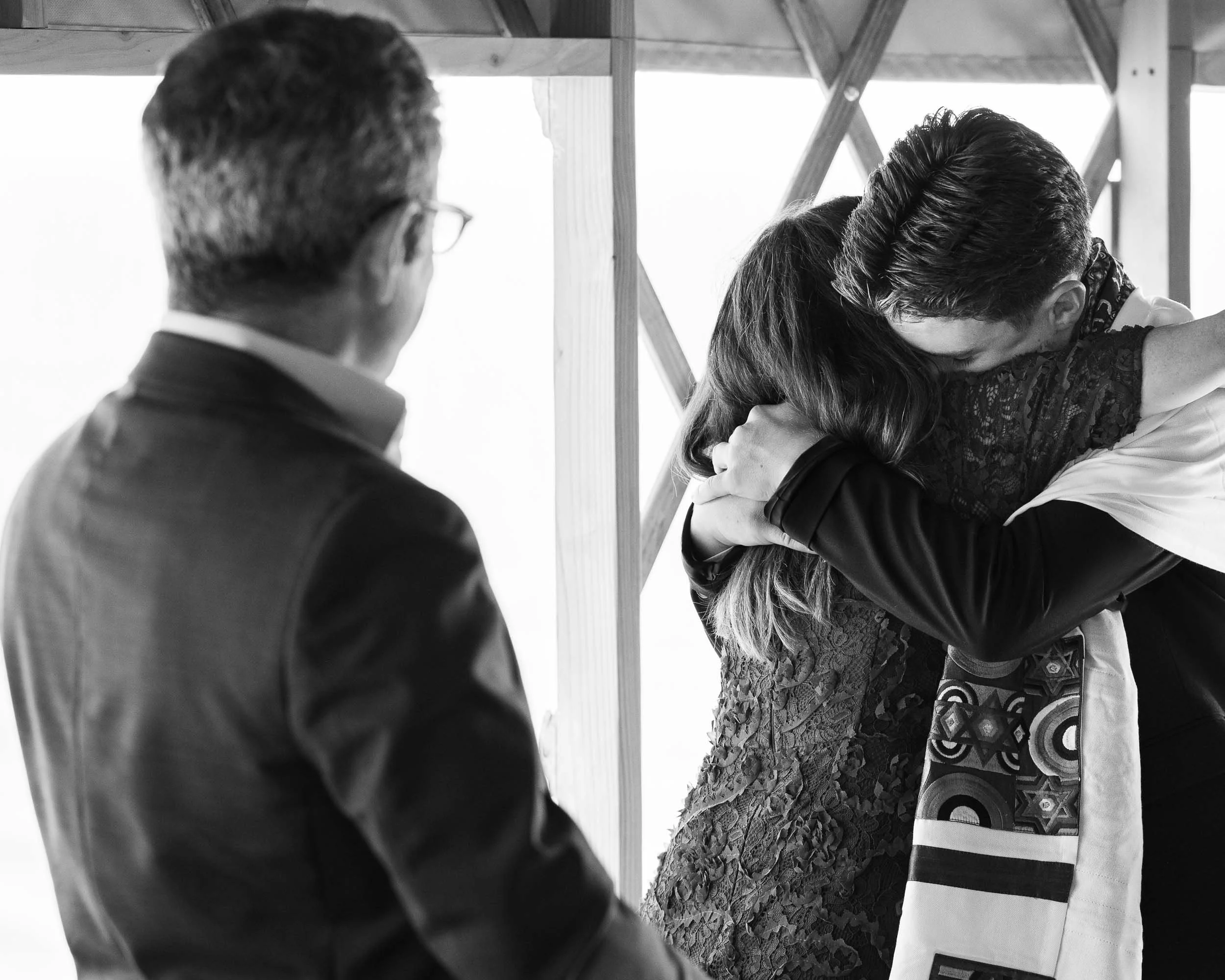 People embracing during a ceremony with an officiant observing, in a setting with wooden beams.