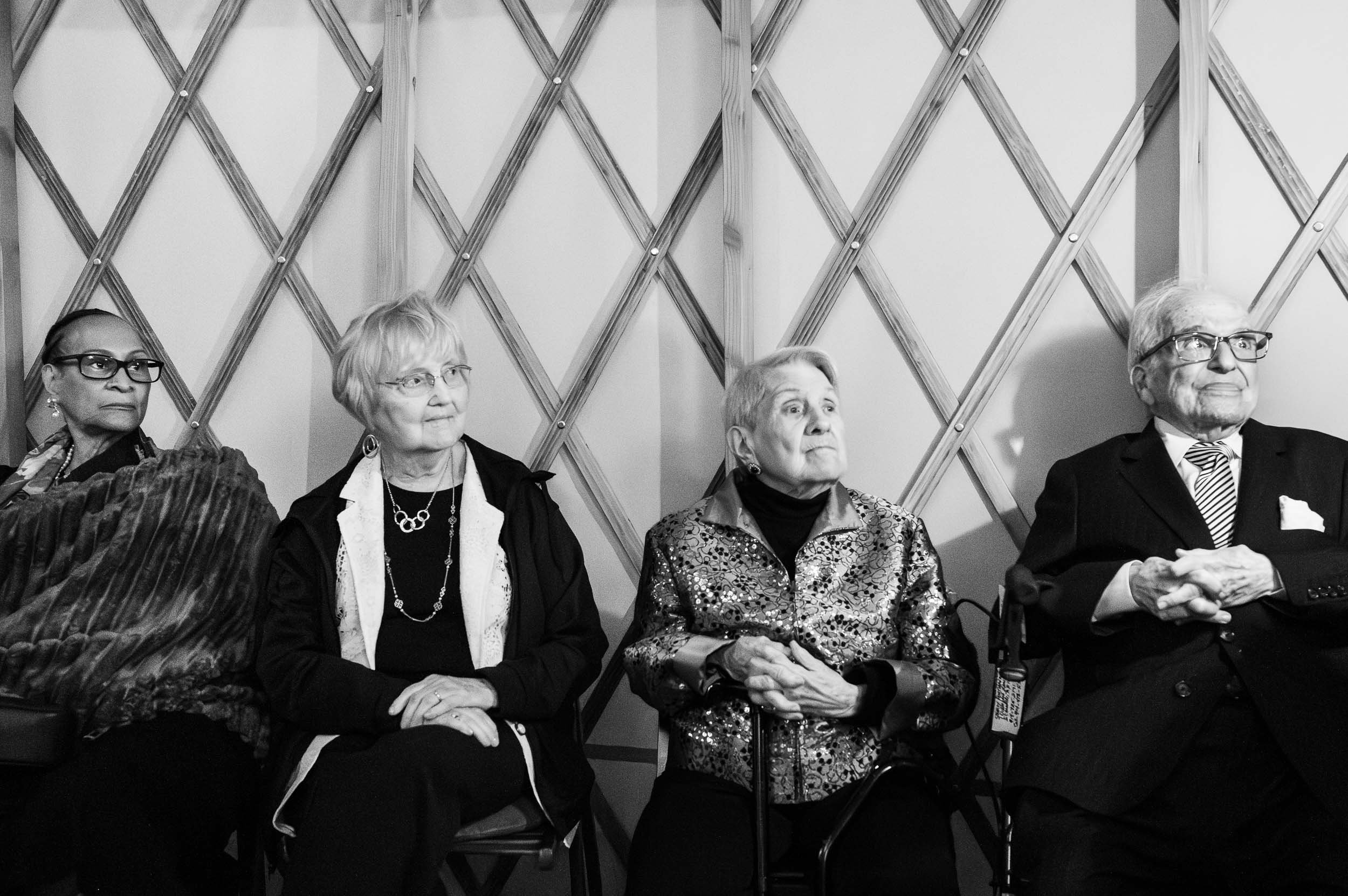 Four elderly adults sitting in chairs against a wooden lattice wall, attentively listening in a black and white photo.