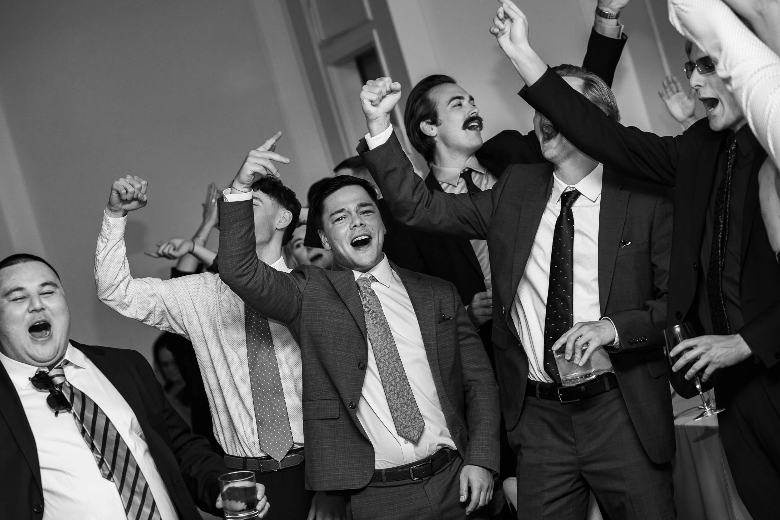 Group of men at a celebration, some with drinks, wearing suits, raising their hands, singing, smiling, enjoying a joyful moment in a social gathering.
