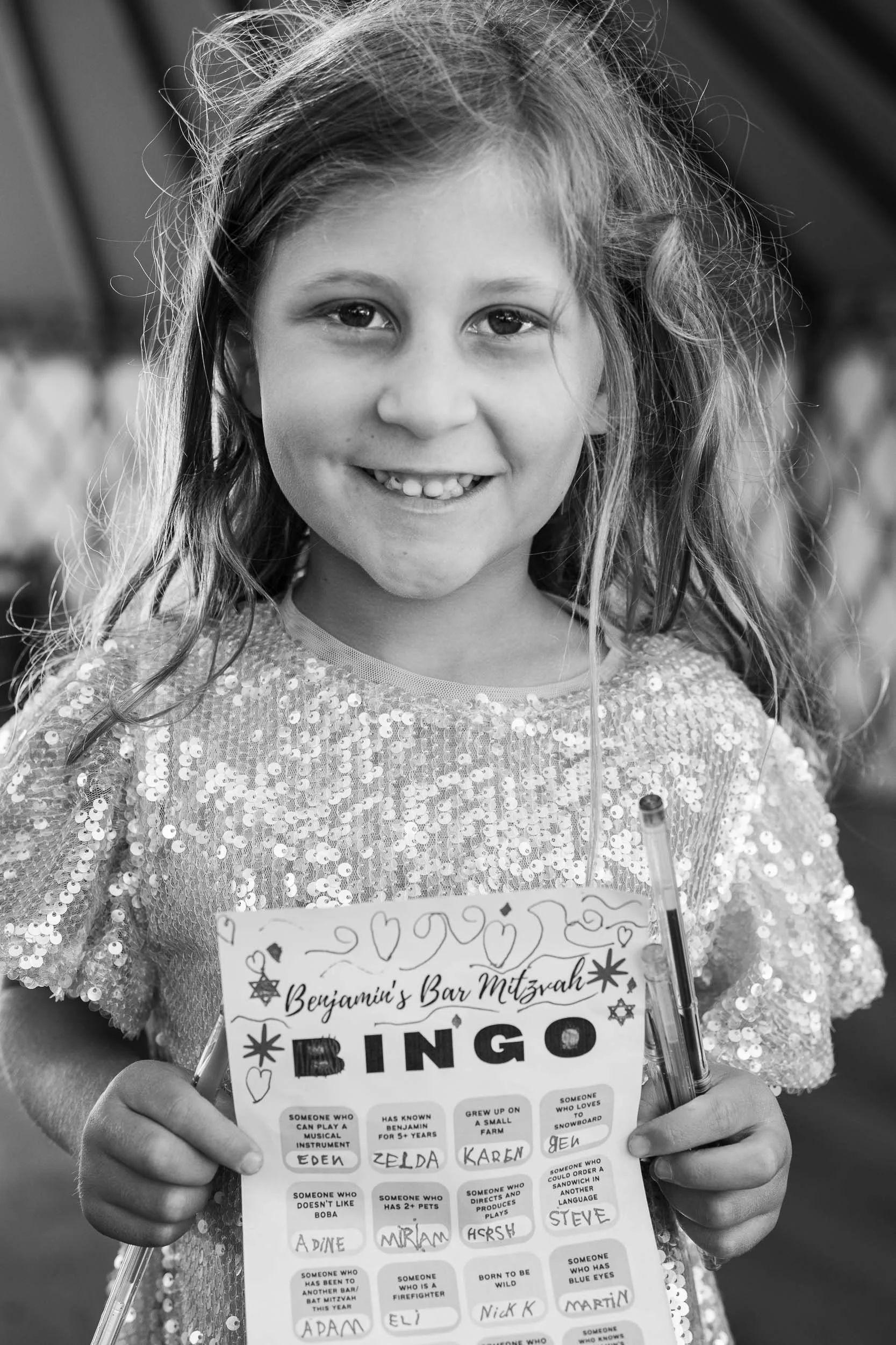 A young girl smiling and holding a bingo card and a pen, wearing a sparkly top, in black and white.