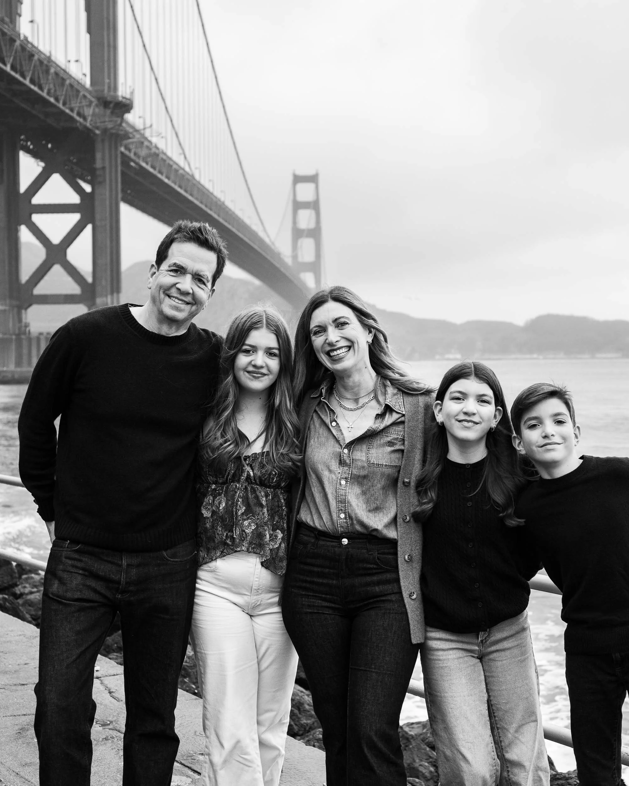 A family of five standing outdoors, smiling, with the Golden Gate Bridge in the background.