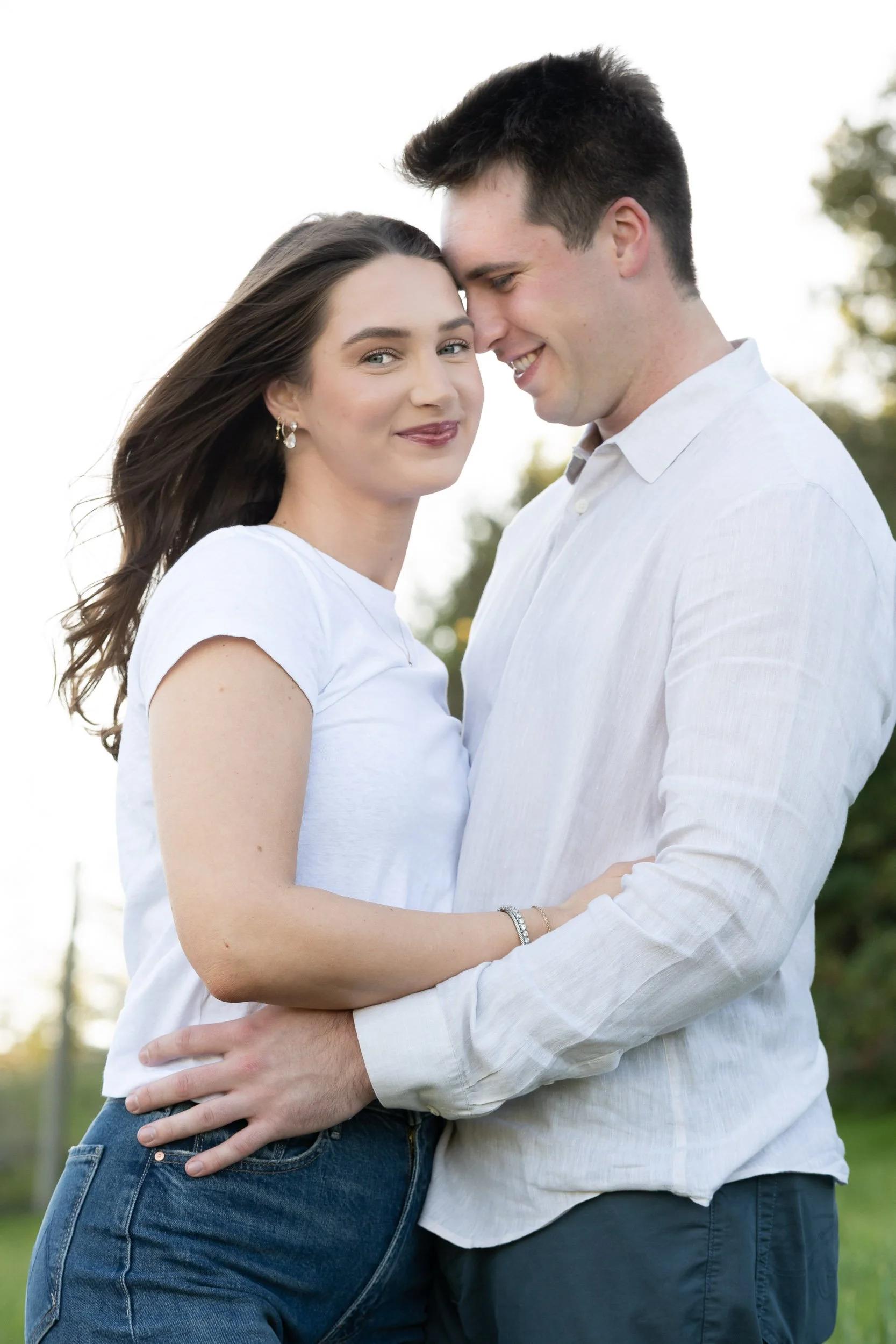 A young couple standing outdoors, holding each other close, smiling, with trees and a bright sky in the background.