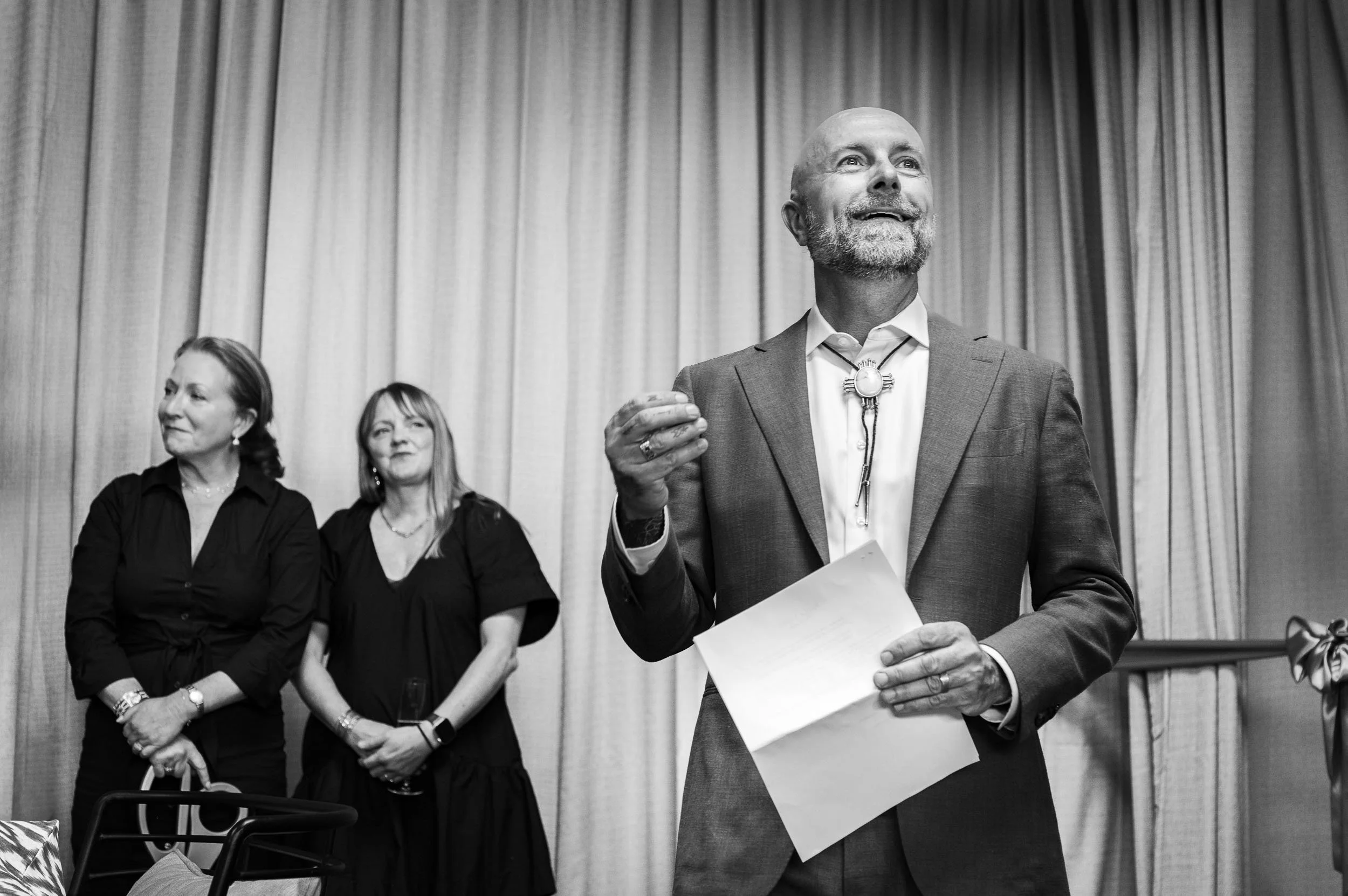 A man in a suit holding papers and gesturing, standing in front of a curtain with two women behind him, all appearing at a formal event.