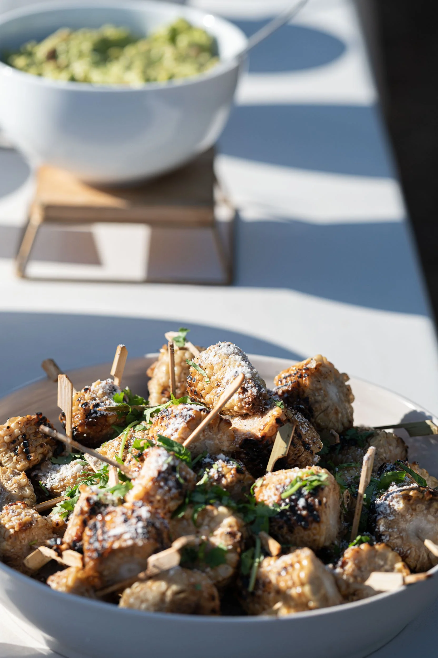 Bowl of grilled chicken drumsticks topped with grated cheese and garnished with fresh herbs, placed on a white table with a bowl of guacamole in the background.