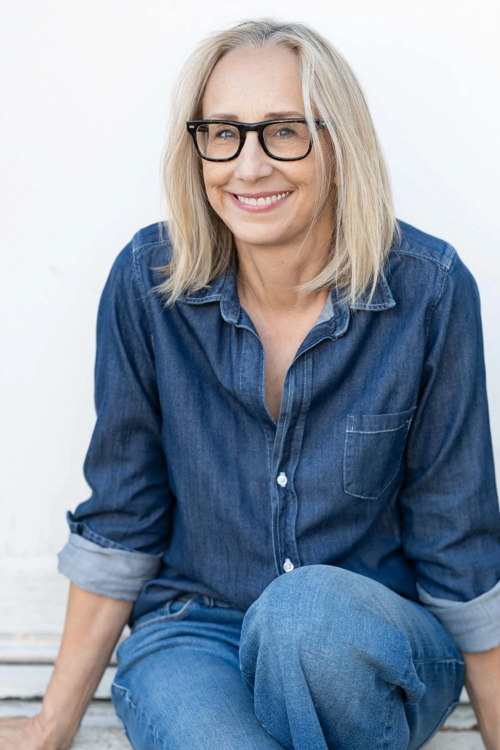 A smiling middle-aged woman with blonde hair, wearing black glasses and a blue denim shirt, sitting on a bench against a white background.