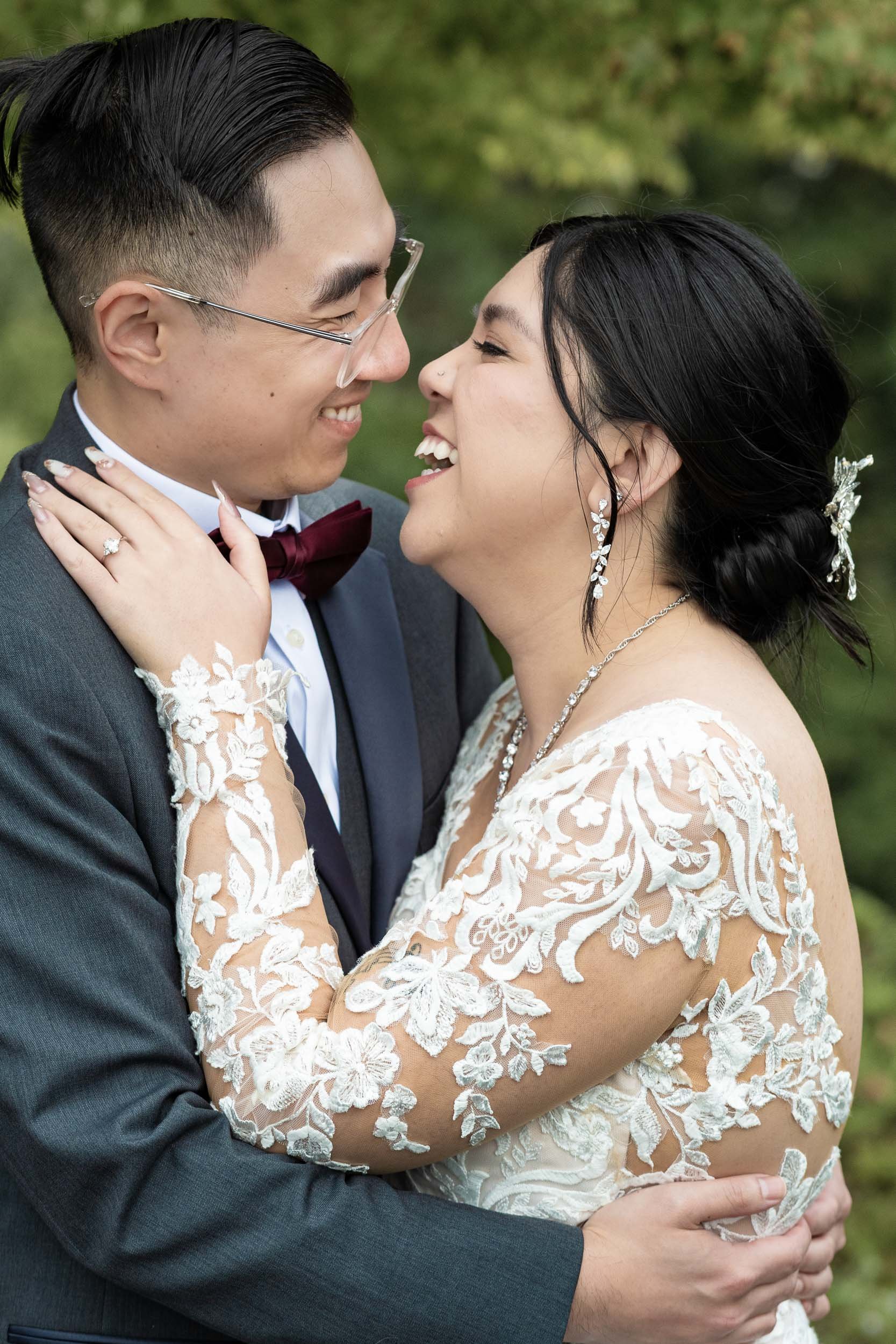 A newlywed couple smiling and embracing each other outdoors, dressed in wedding attire with a lush green background.