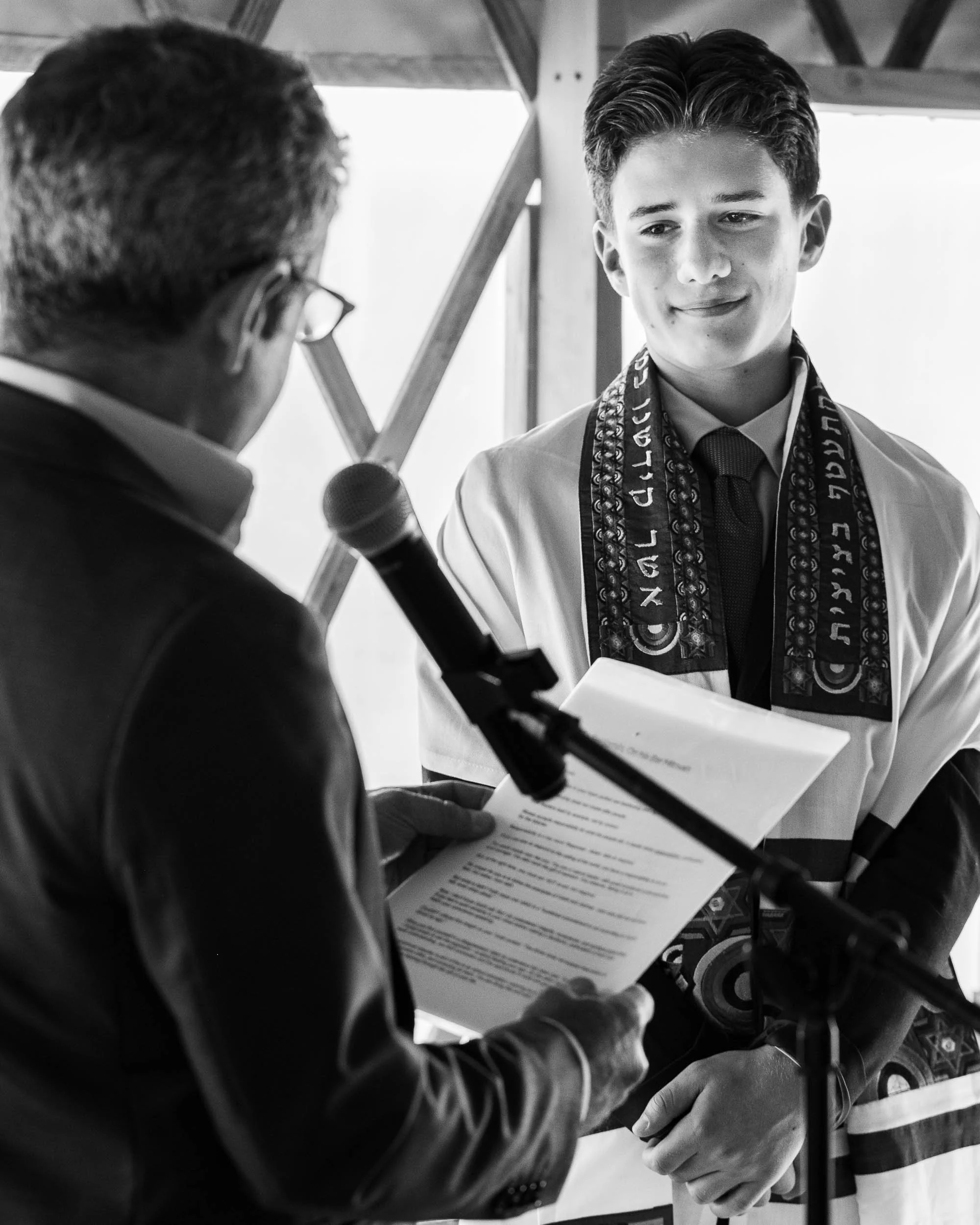 A young man receiving an award or recognition, wearing a sash with decorative patterns, as an older man holds a certificate and speaks into a microphone.