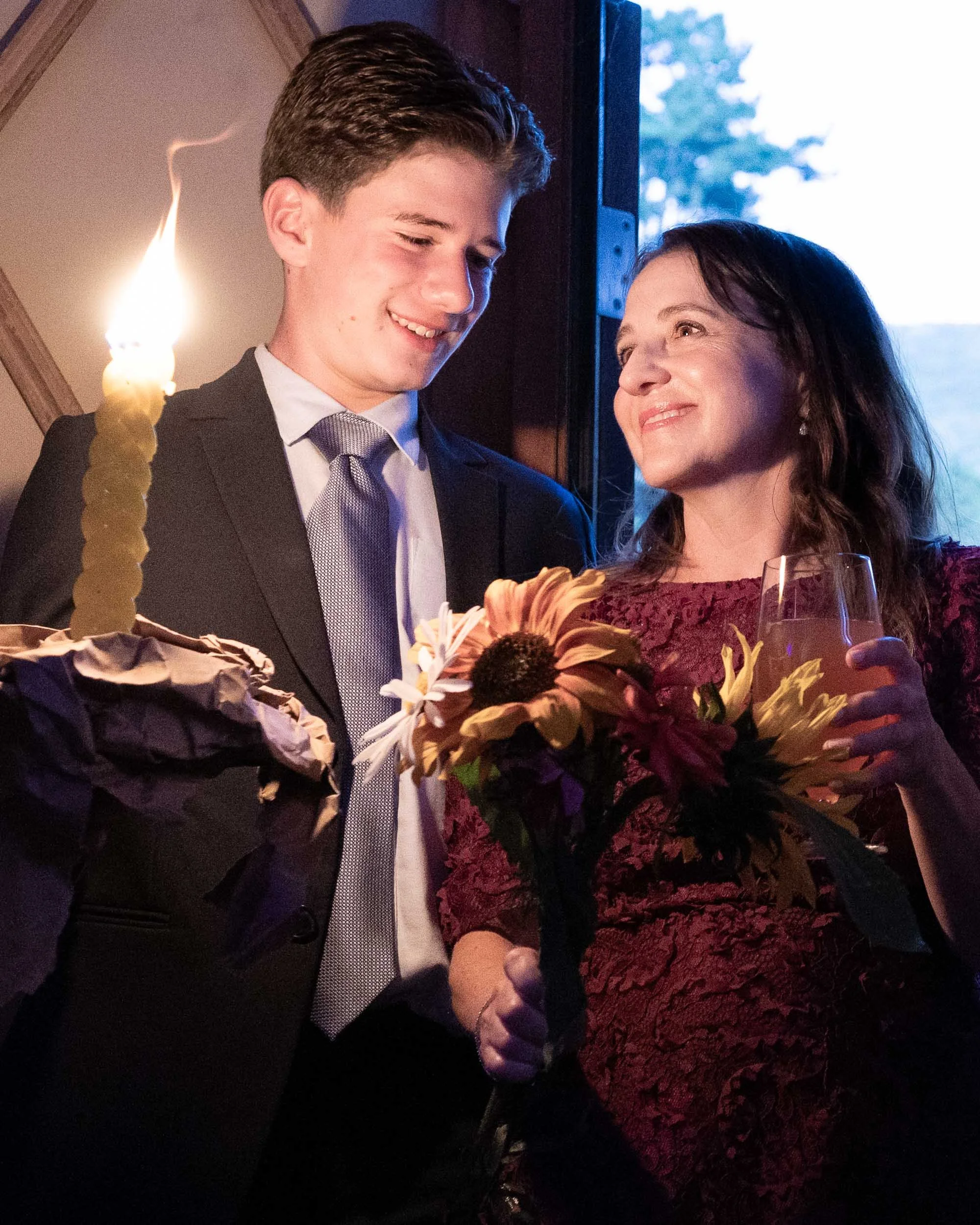 A young man in a suit celebrates with a woman holding flowers and a wine glass, near a birthday cake with a lit candle.