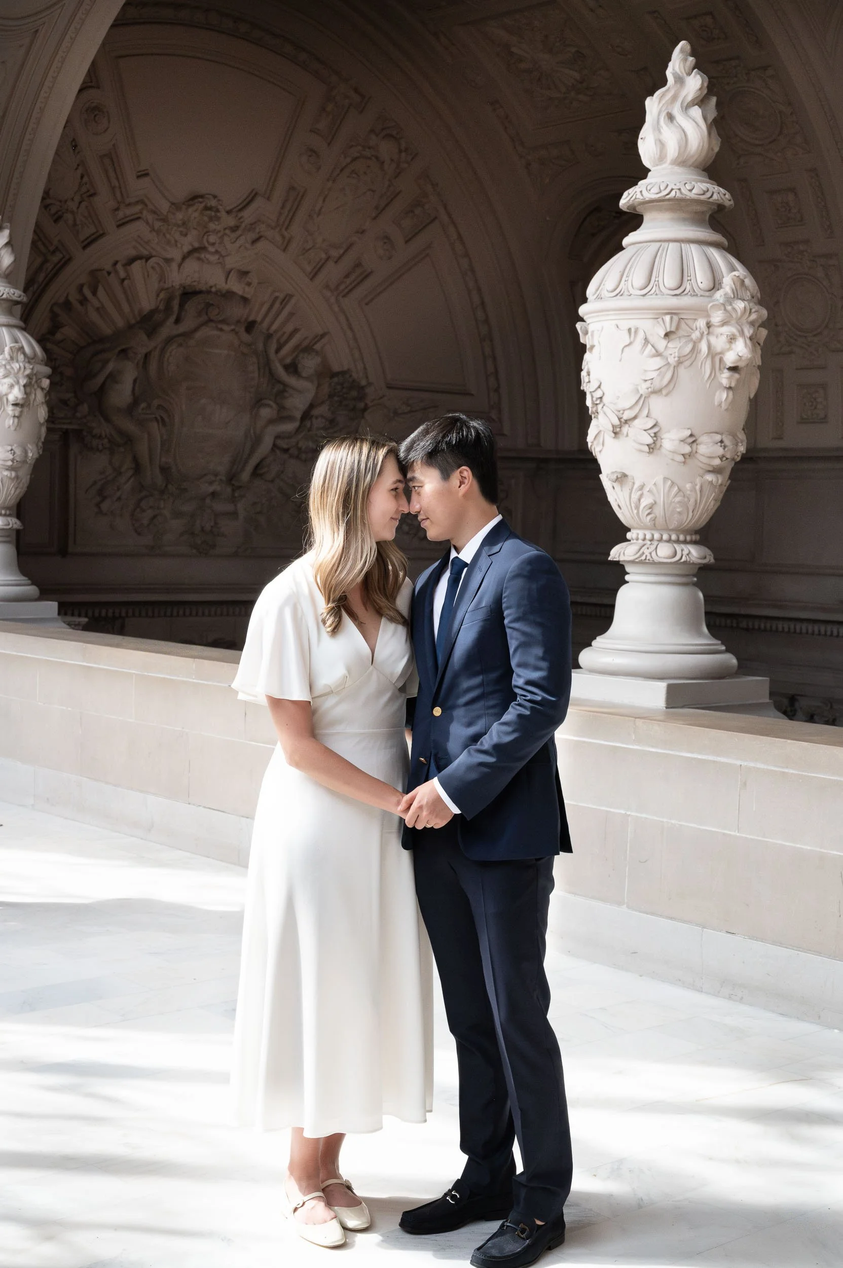 A couple dressed in formal attire standing close together in a grand, ornate hallway with classical architectural details and large decorative vases.