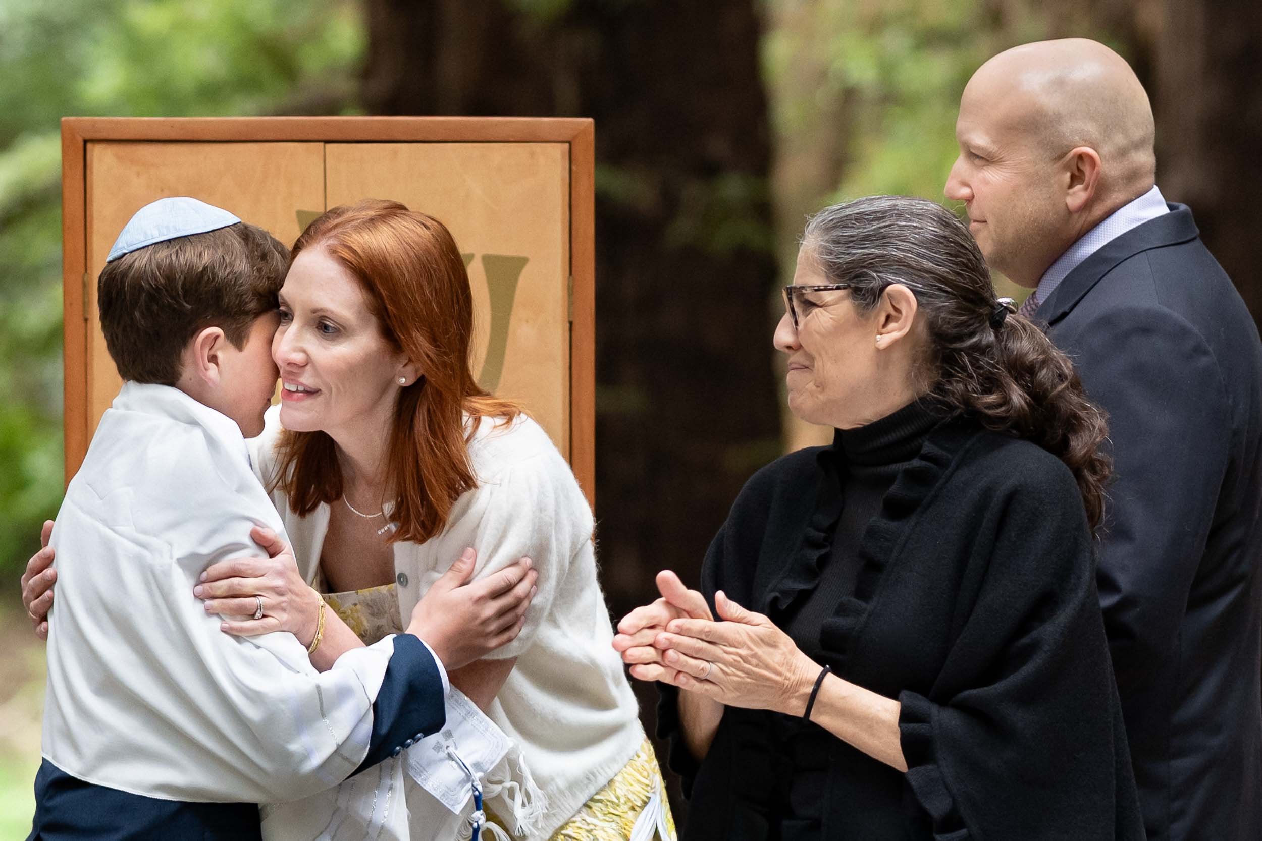 A group of five people, including a woman with red hair and glasses, a man with glasses and a beard, a woman with gray hair, a man with a yellow kippah, and a woman's child, are engaged in a meaningful embrace and conversation outdoors against a blur
