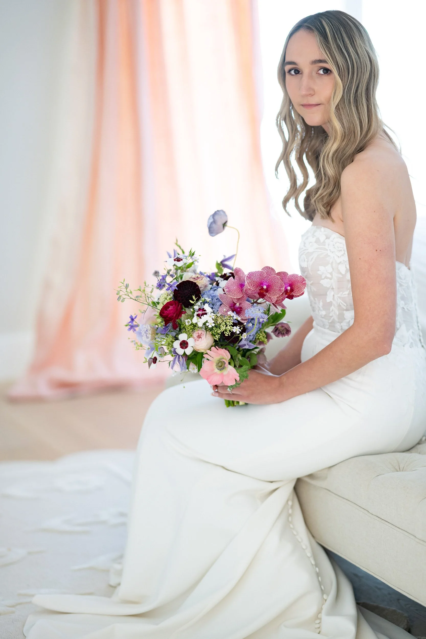 A woman in a white strapless wedding dress, holding a colorful bouquet of flowers, sitting on a white chair in a softly lit room with curtains in the background.