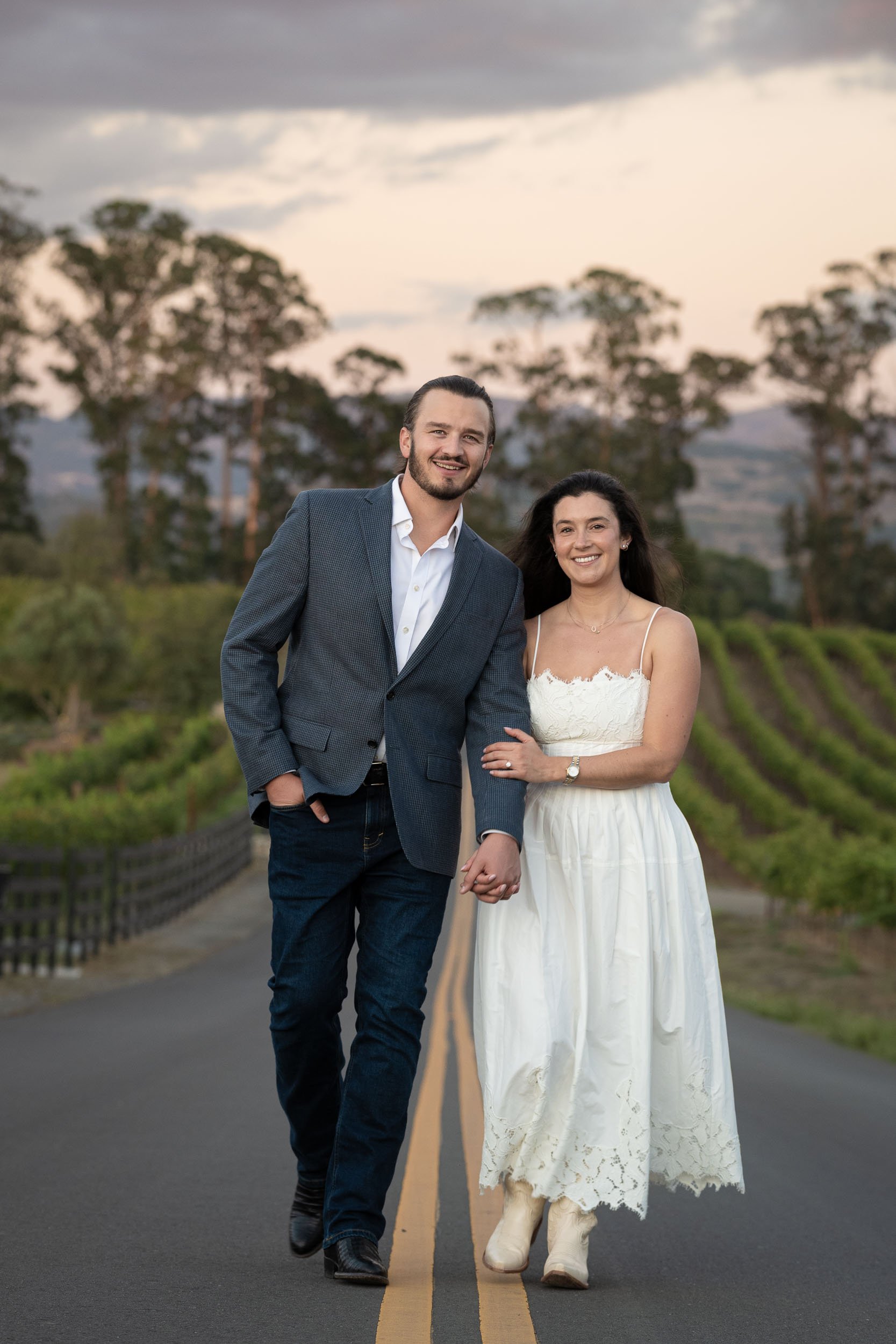A happy couple walking hand in hand down a rural road at sunset, with a green vineyard and tall trees in the background.
