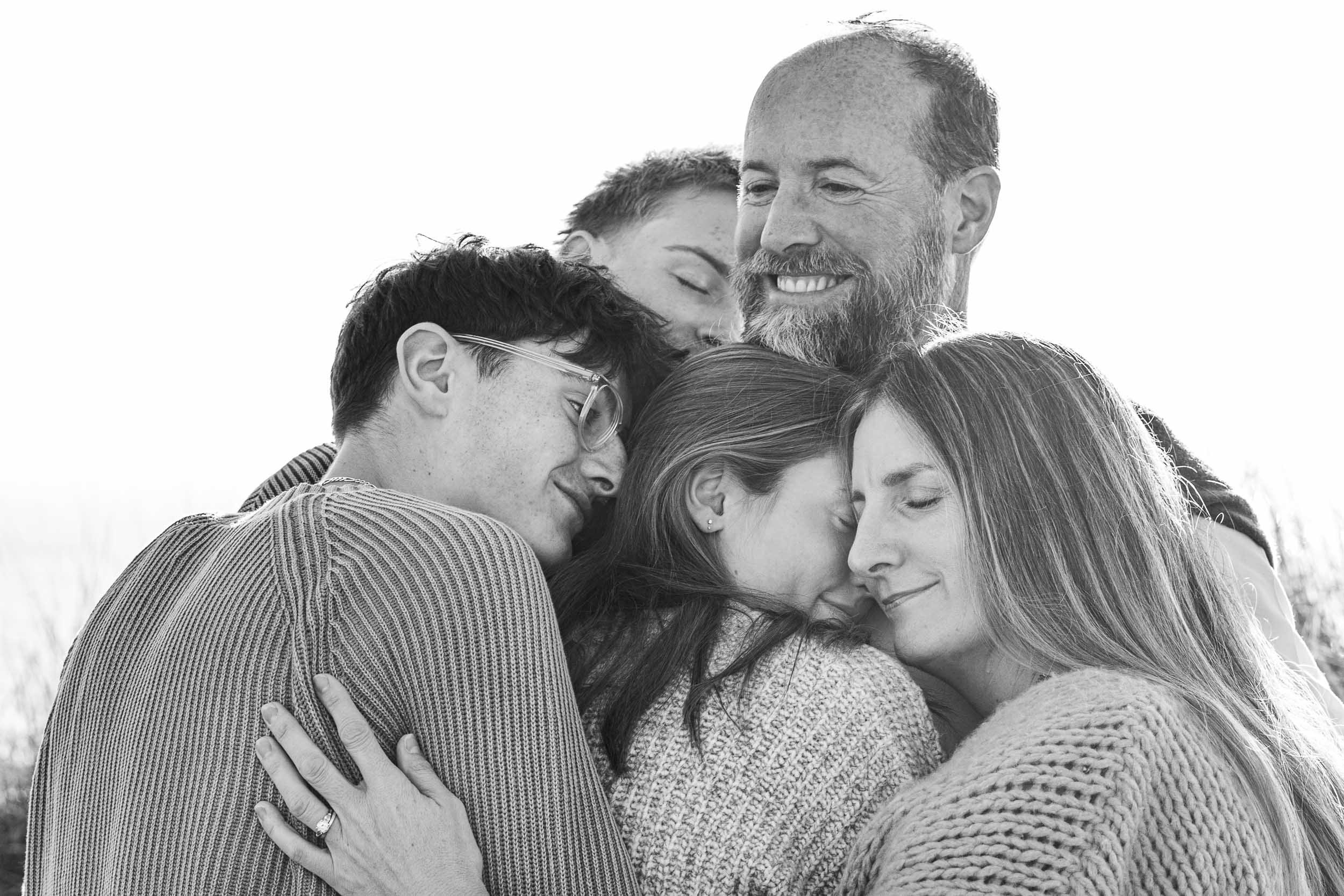 Black and white photograph of six people hugging each other outdoors, eyes closed and smiling, showing close bond and affection.