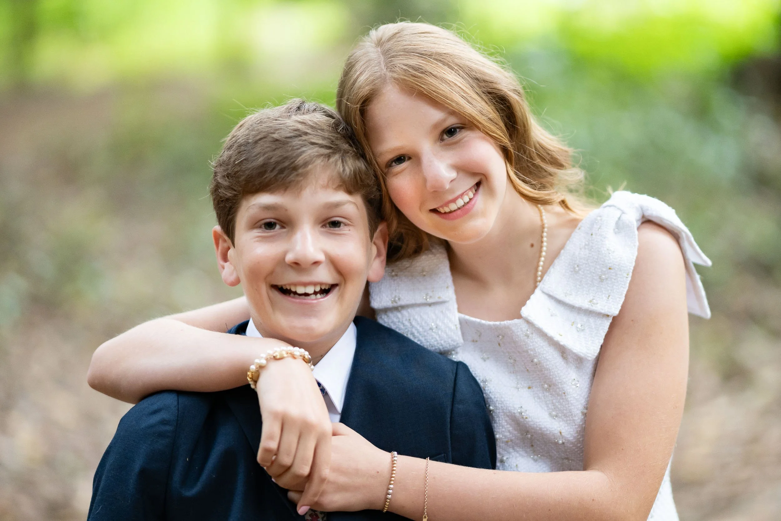 A young woman and a young man are smiling and hugging outdoors with a blurred green background.
