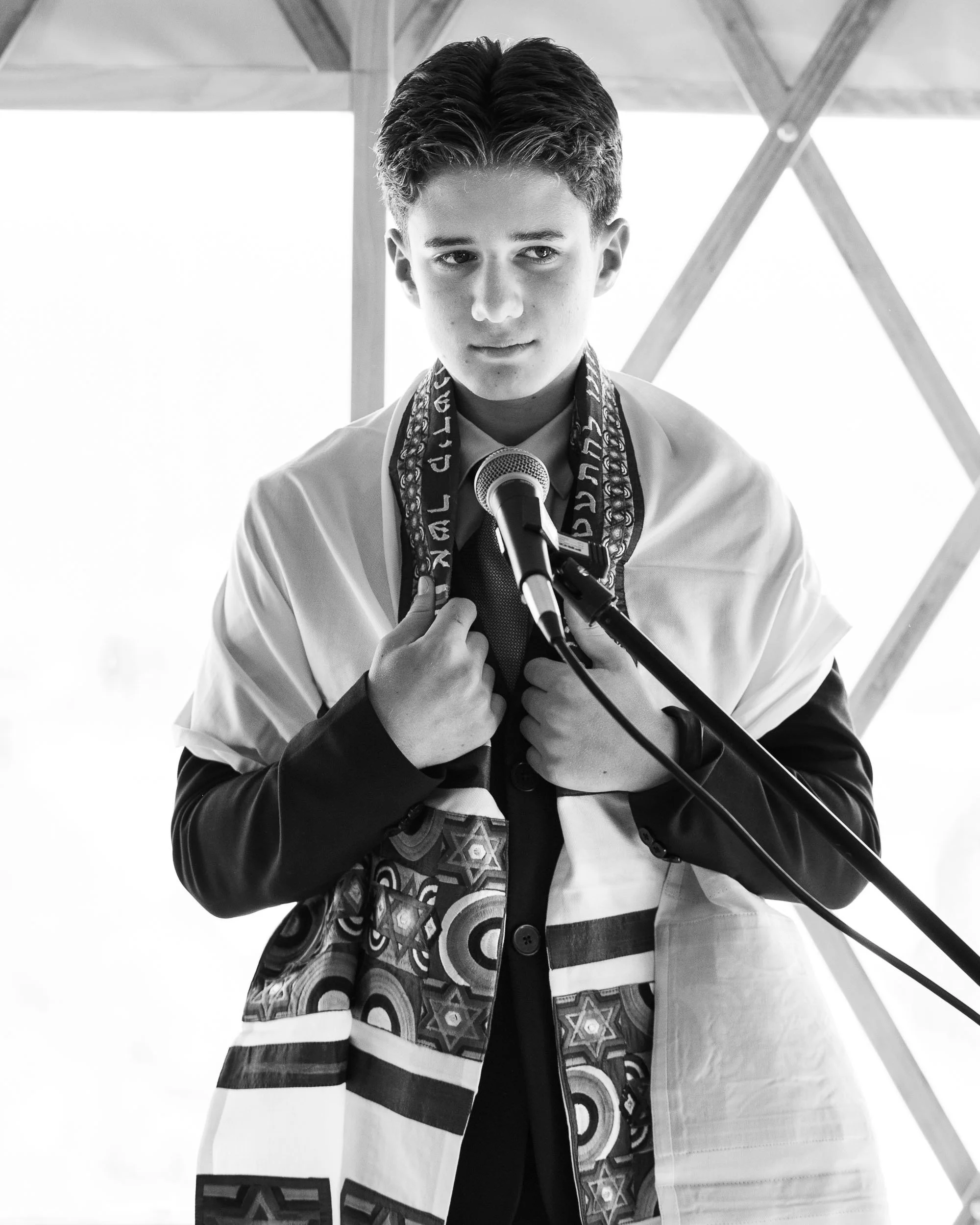 Young man at a microphone, adjusting a ceremonial shawl around his shoulders, in a setting with wooden beams in the background.