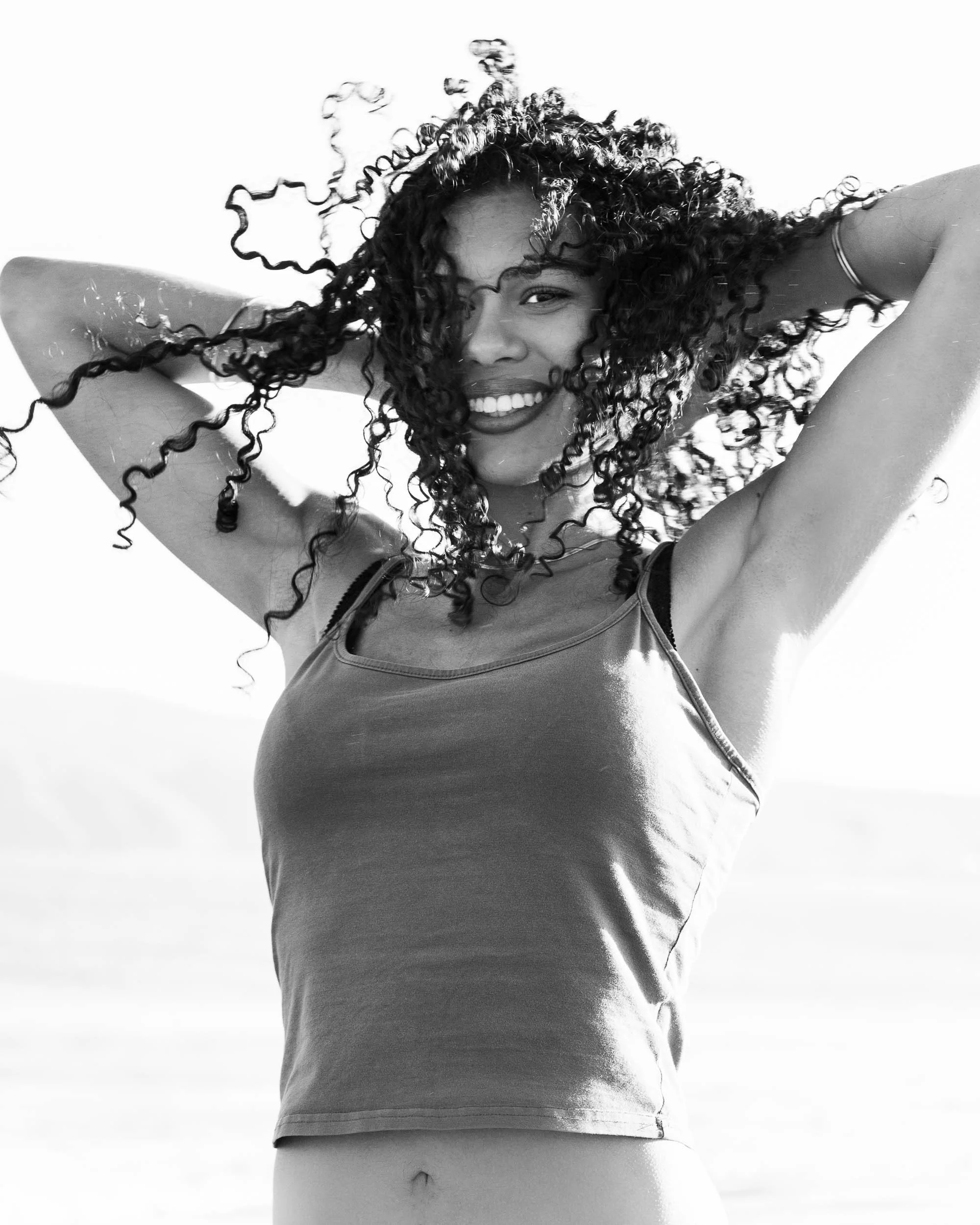 A woman smiling with curly hair, raising her arms, and posing outdoors.