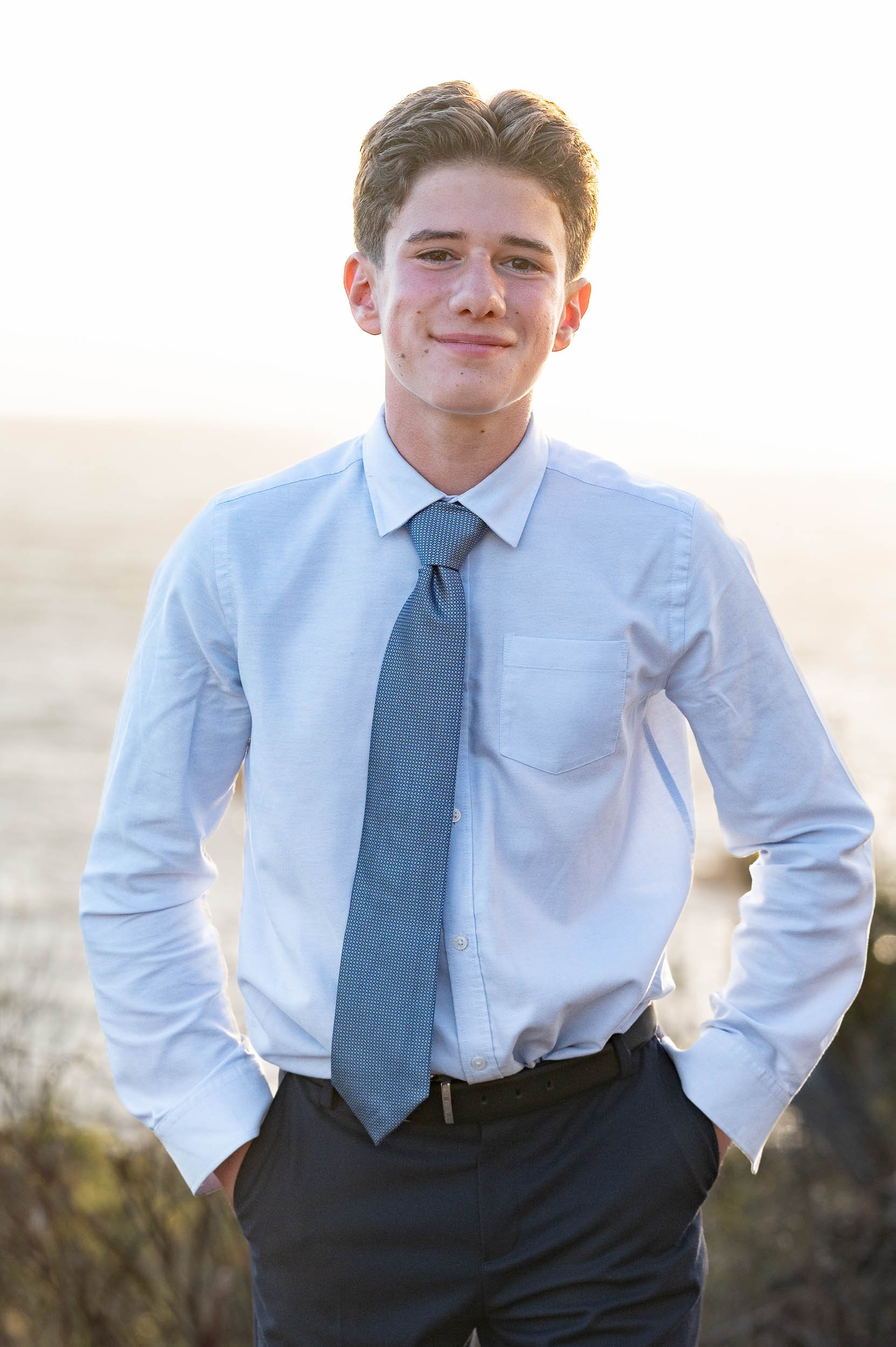 A young man standing outdoors in a light blue dress shirt and tie, with hands in his pockets, smiling at the camera, in front of a blurred natural landscape during sunset.