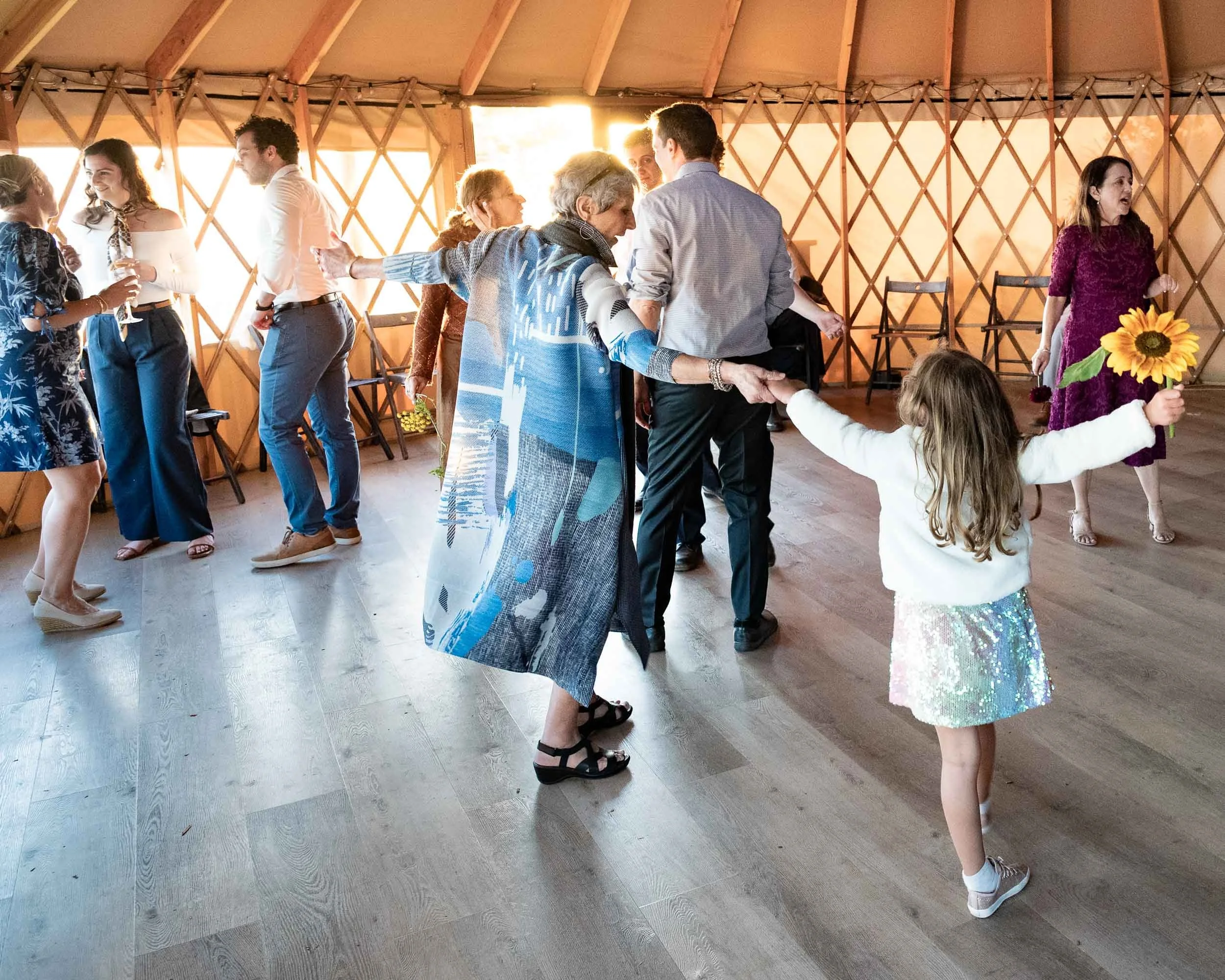 People dancing and socializing inside a wooden structure during a celebration, with a young girl holding a sunflower and adults holding hands.