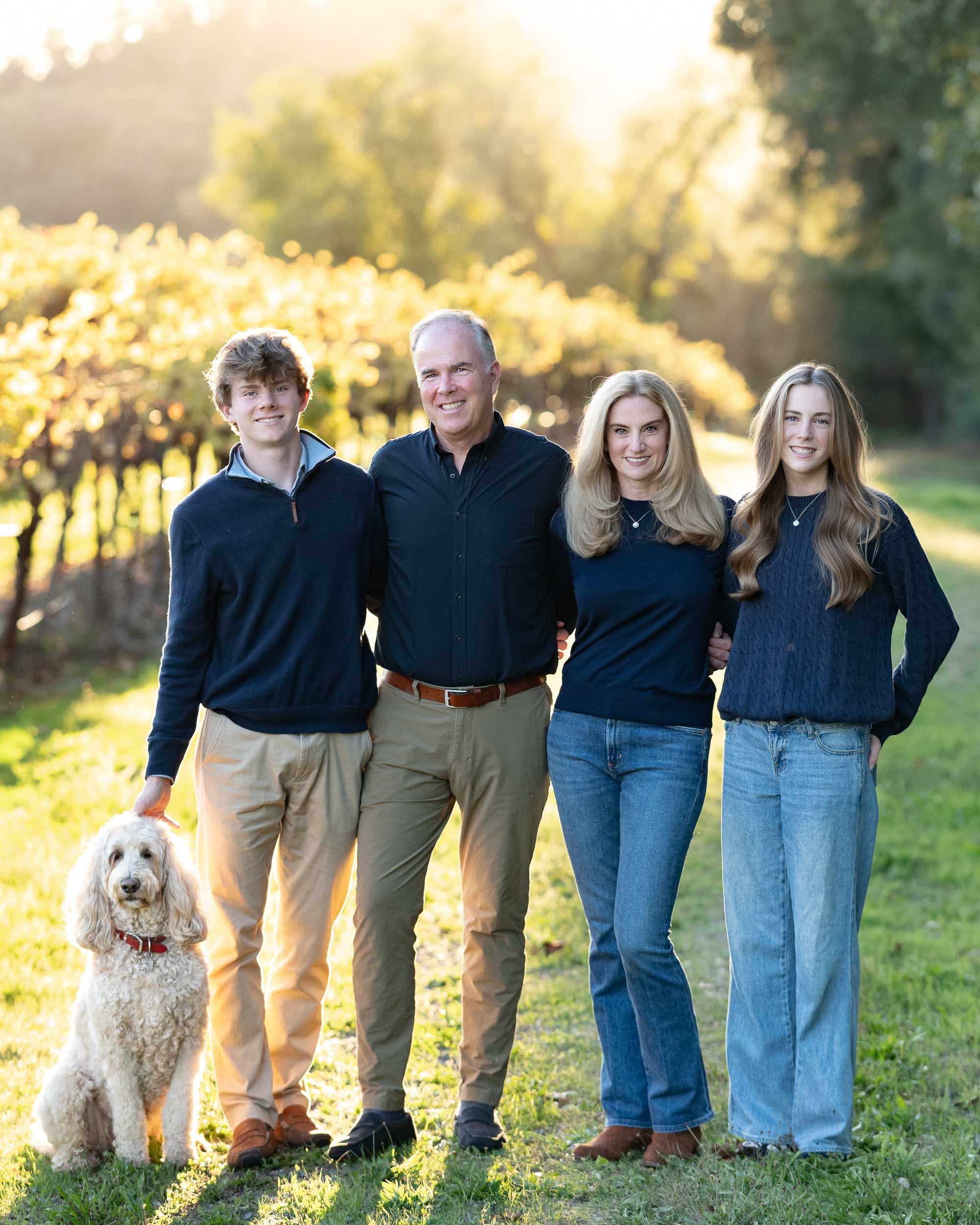 Family of four and dog standing outdoors in autumn sunlight, smiling together.