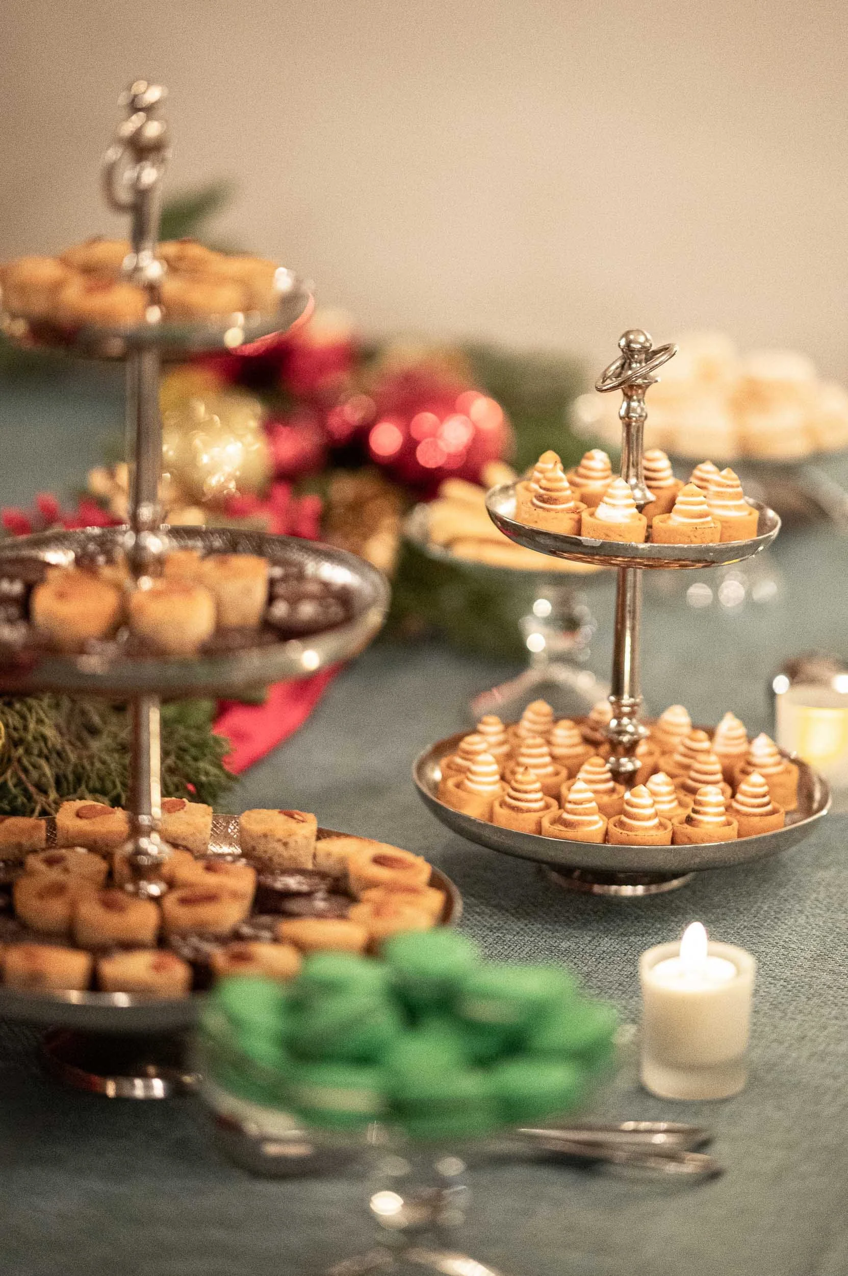 Tiered serving trays filled with assorted cookies and confections, decorated for a festive holiday celebration, with candles and holiday decorations in the background.