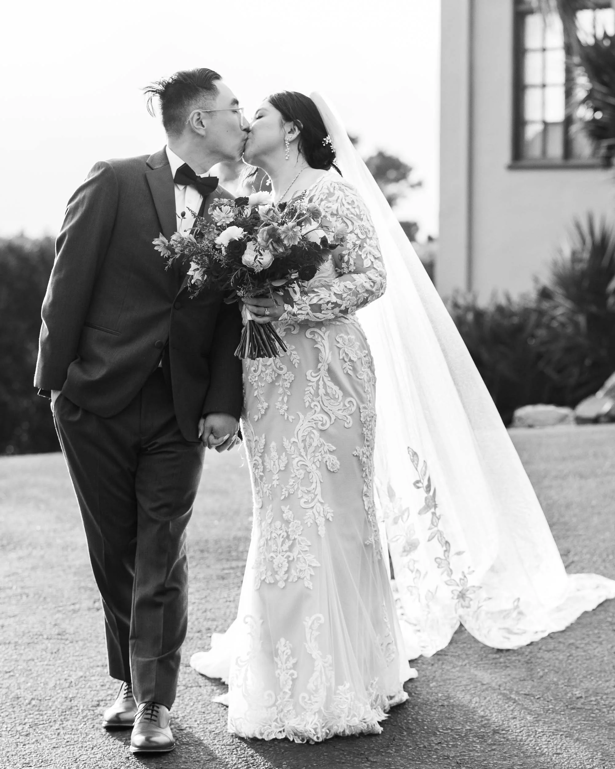 A bride and groom share a kiss while holding hands outdoors during their wedding. The bride is in a lace wedding gown and veil, holding a bouquet, and the groom is dressed in a suit with a bow tie.