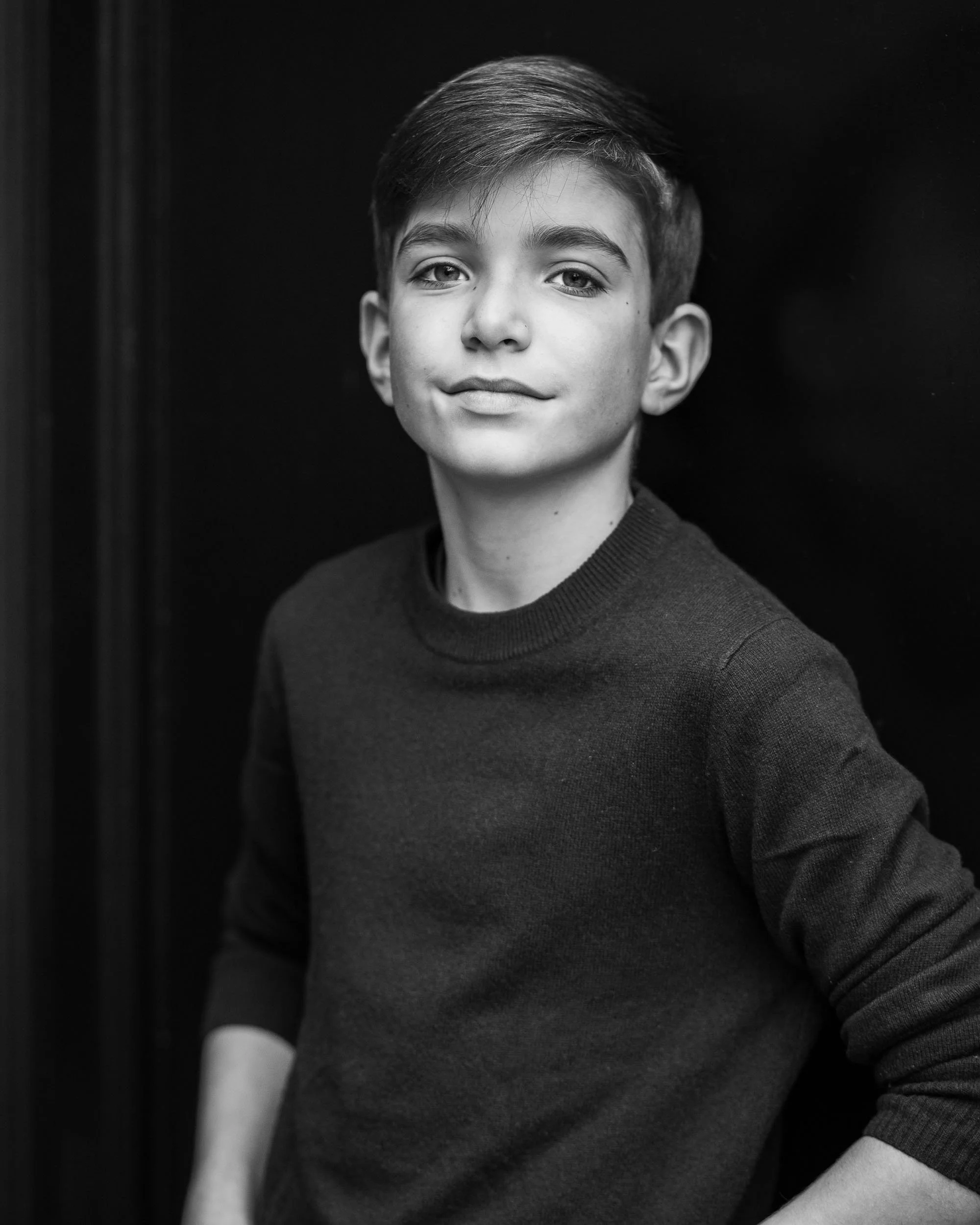 Black and white portrait of a teenage boy with short hair, wearing a dark sweater, standing with arms akimbo against a dark background.