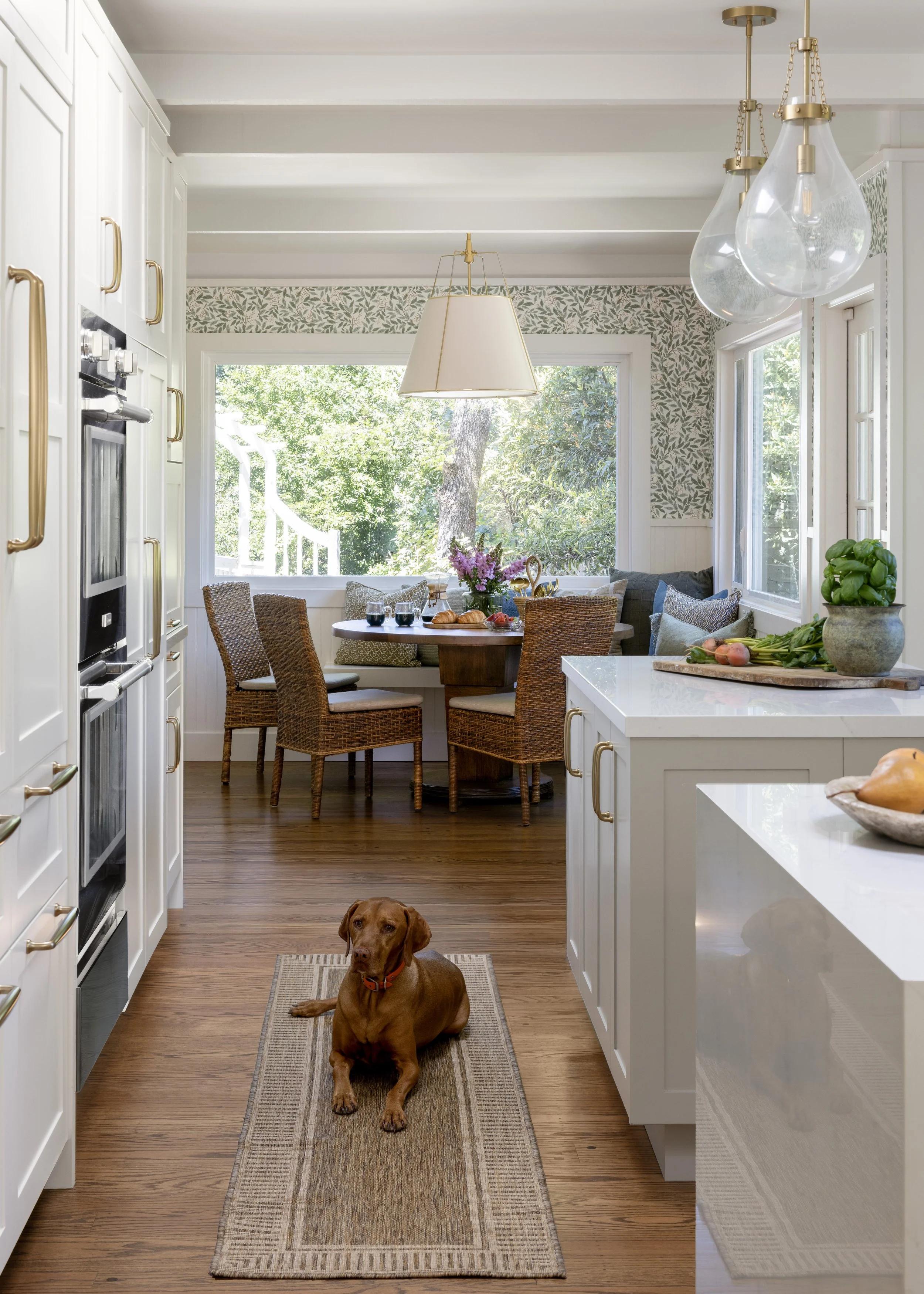 Kitchen renovation in Kentfield designed by Marin interior designer Studio SQUIRE featuring a Window Seat, Walnut Round Table, Pillows, Wicker Chairs, Brass Pendants, Quartz Countertop, Custom Cabinetry, Brass Hardware, and Floral Wallpaper