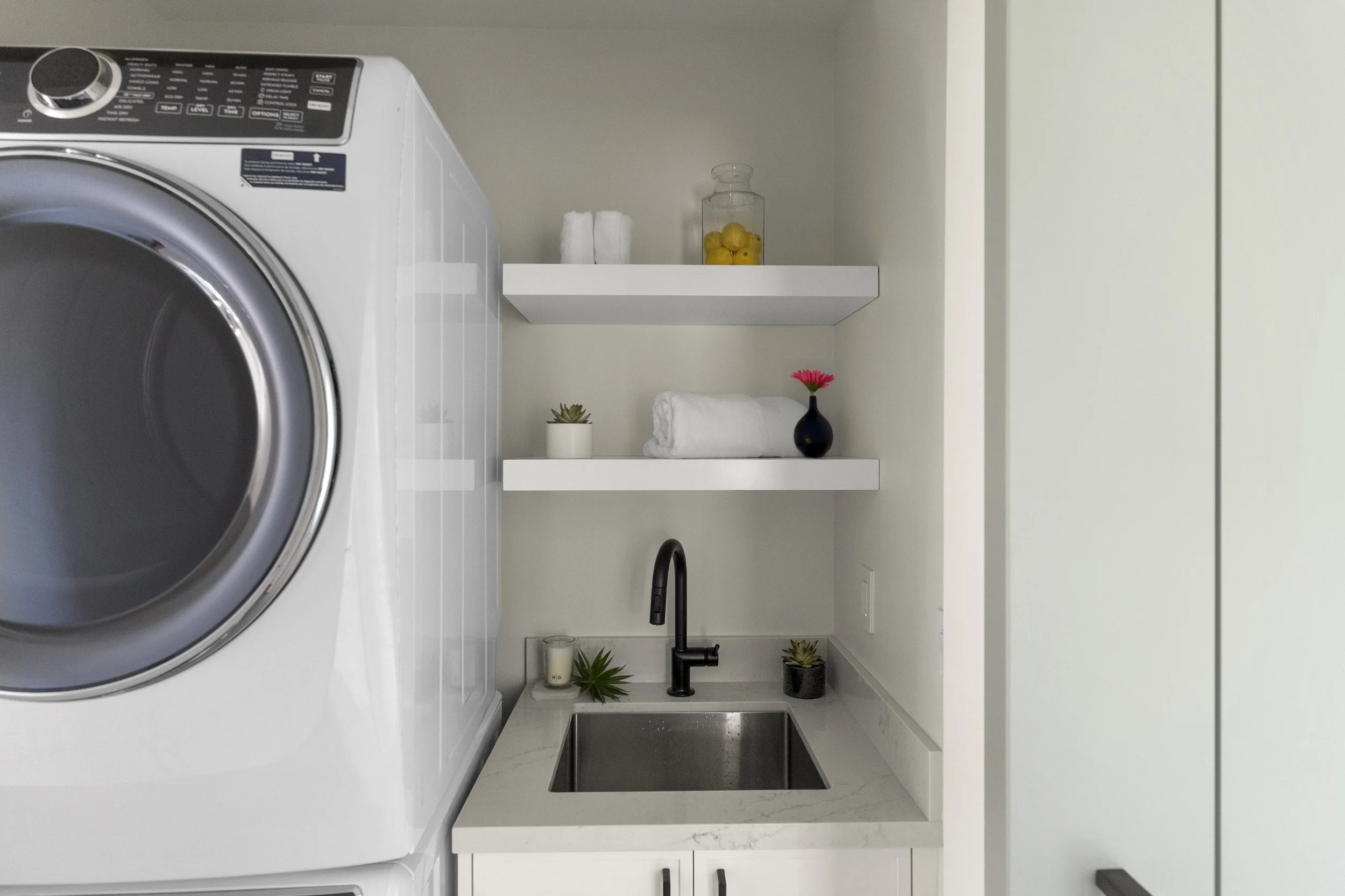 Laundry closet renovation in Tiburon designed by Marin interior designer Studio SQUIRE featuring a sliding barn door, small sink, open shelving, and a stacking washer dryer.