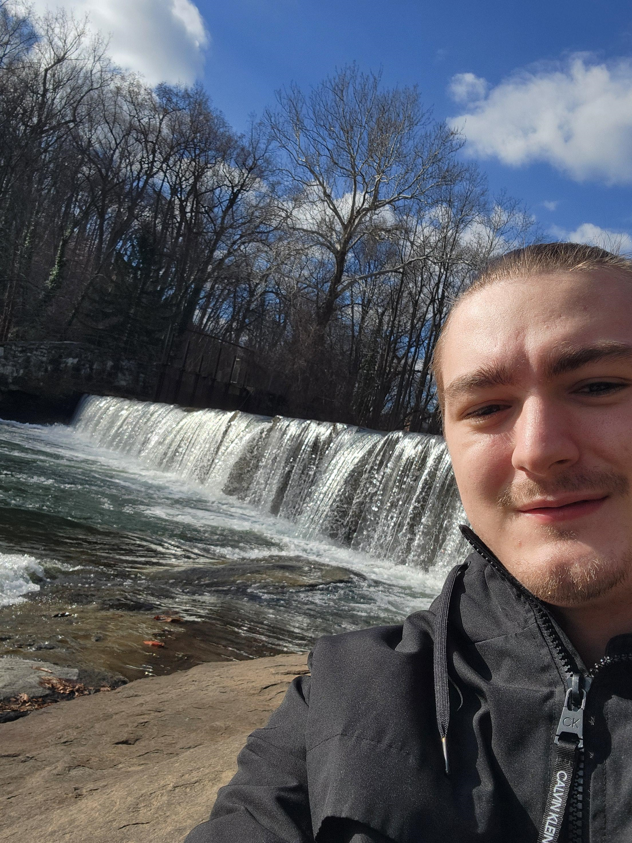 A young man taking a selfie outdoors near a waterfall, with leafless trees and a blue sky with clouds in the background.