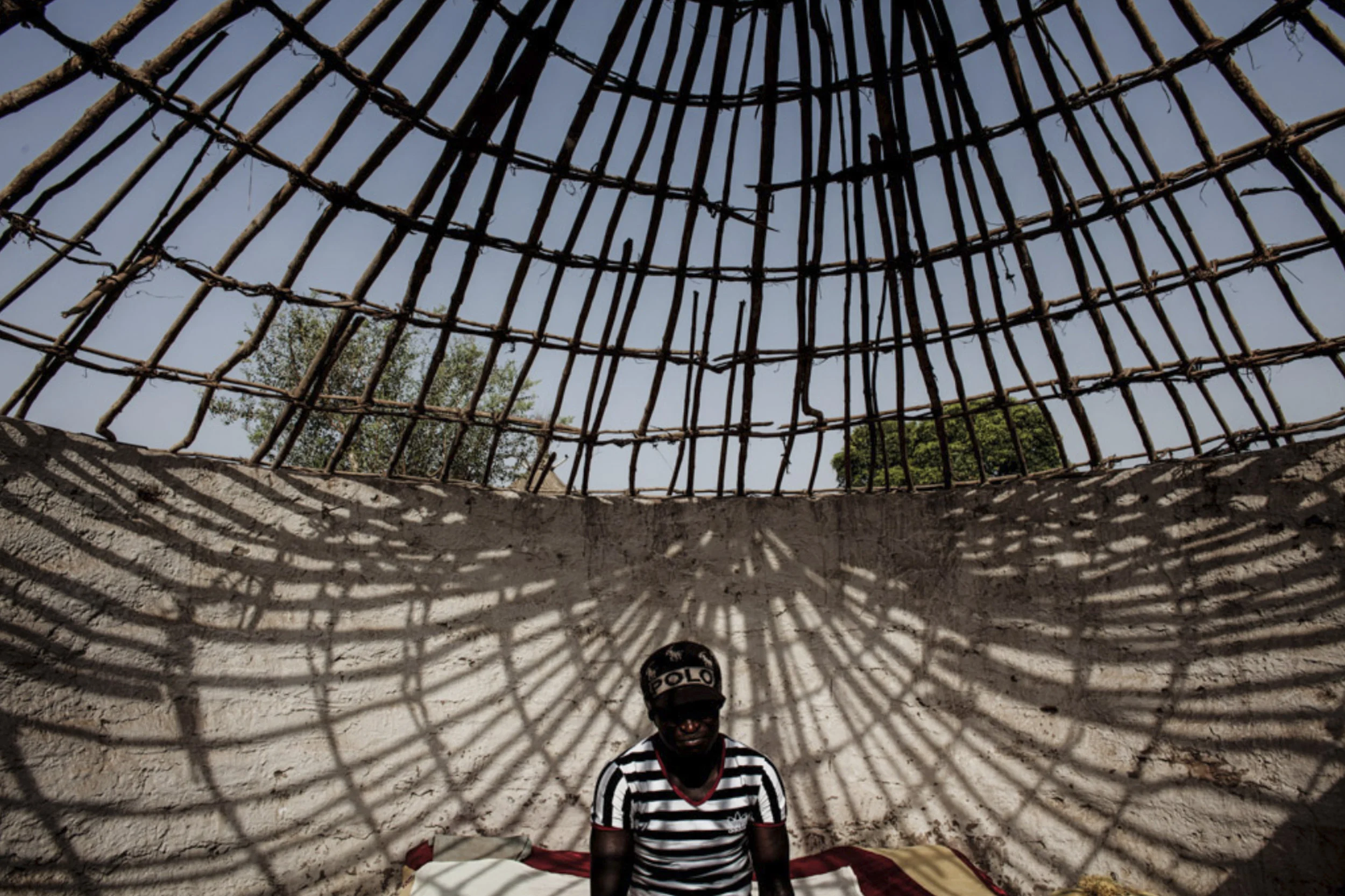 Fissa, 33, sits on his bed under the framing for his thatched roof at his home in Perai Village, Gambia. Fissa, like many other men in his village plans to take the “Back Way” overland from The Gambia, through Senegal, Mali and Burkina Faso then Niger through the desert to Libya across the sea on to Europe hoping to find a way to provide for his immediate and extended family.