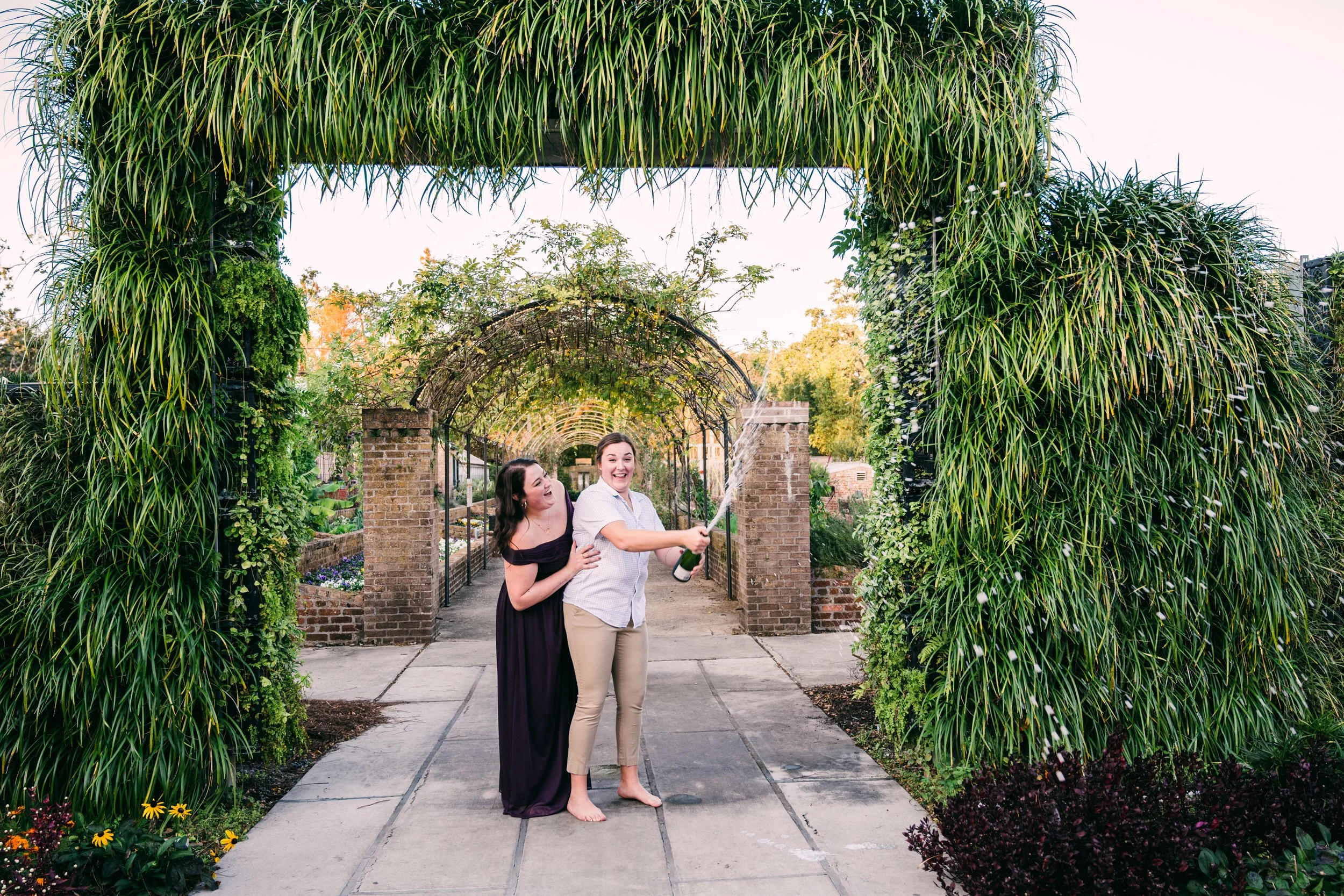 A couple laughing as they stand together and pop a bottle of champagne