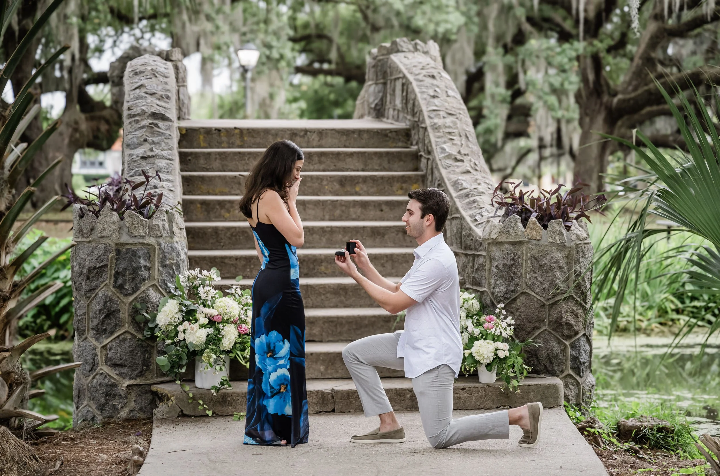 A person kneeling down proposing to their partners in front of a stone bridge