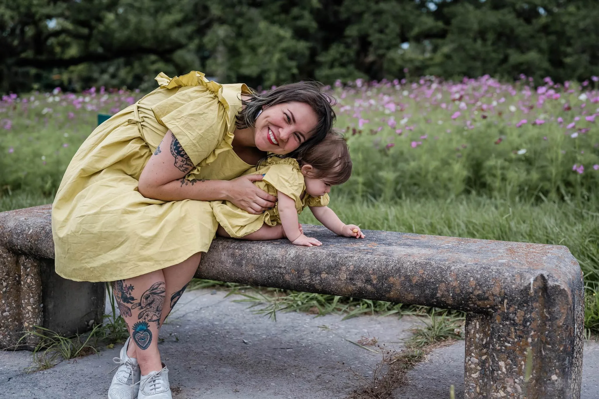 Ash leaning over her child on a stone bench as she smiles