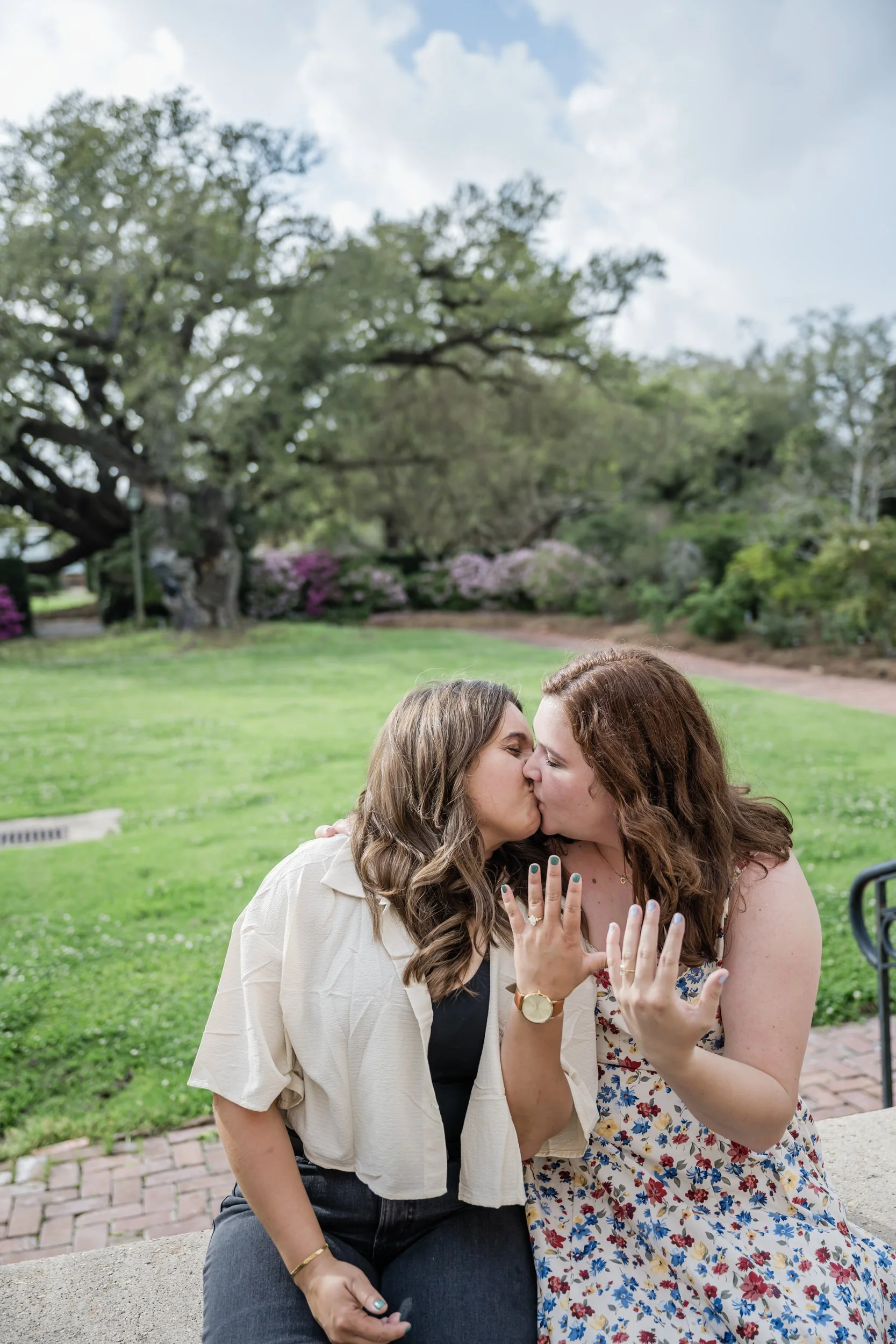 A couple sitting next to each other kissing as they both show off engagement rings