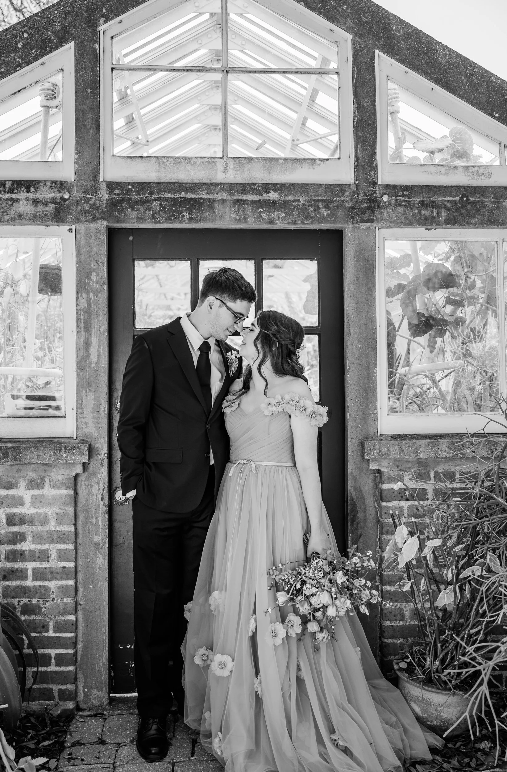 A newlywed couple standing outside of a greenhouse as they lean in to kiss