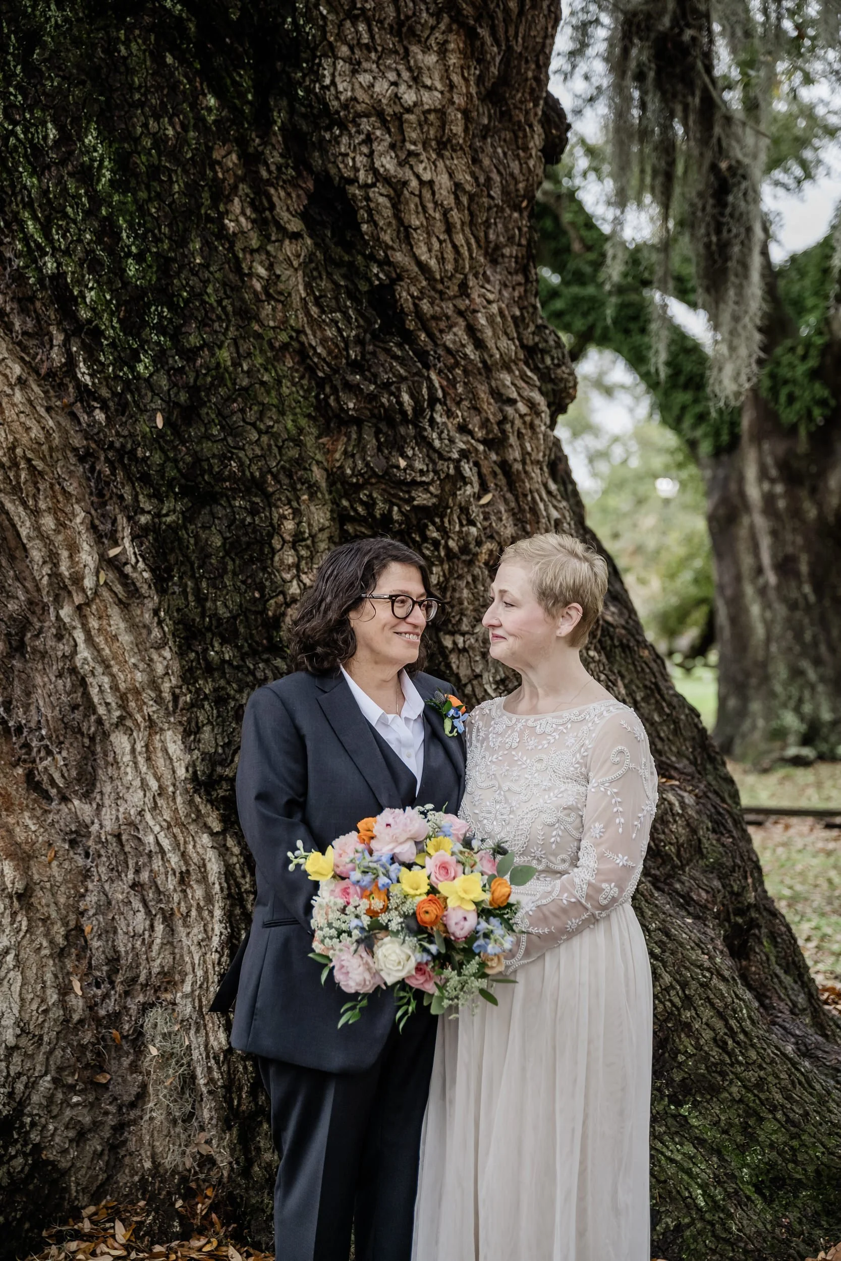 Newlyweds smiling at each other as they stand next to a tree