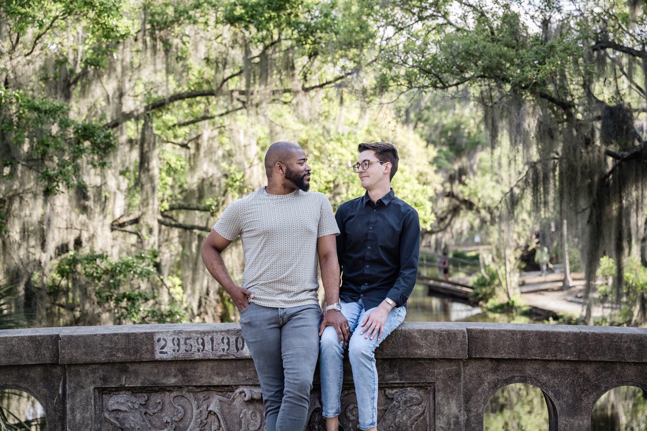A couple smiling at each other as one sits on the edge of a small bridge and the other stands next to them