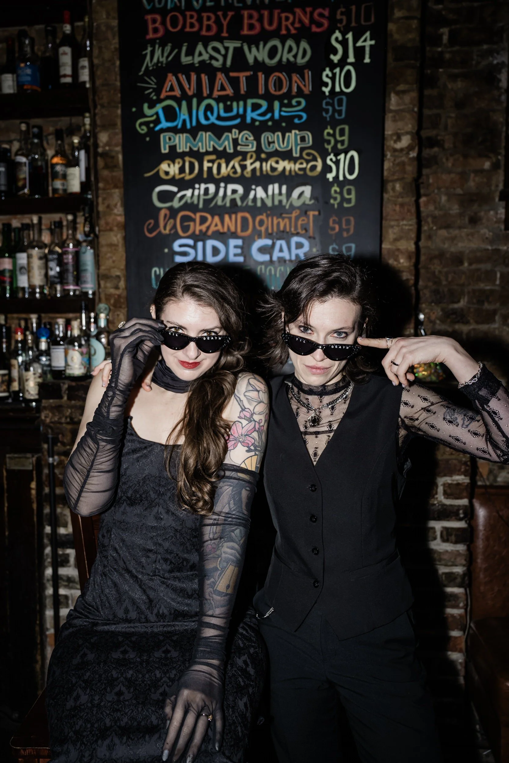 A couple standing against a wall both looking over the top of lowered sunglasses