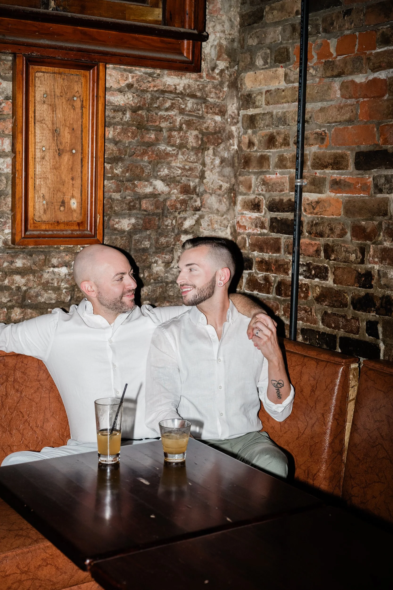 A couple sitting in a corner booth smiling as one has their arm around the other's shoulder