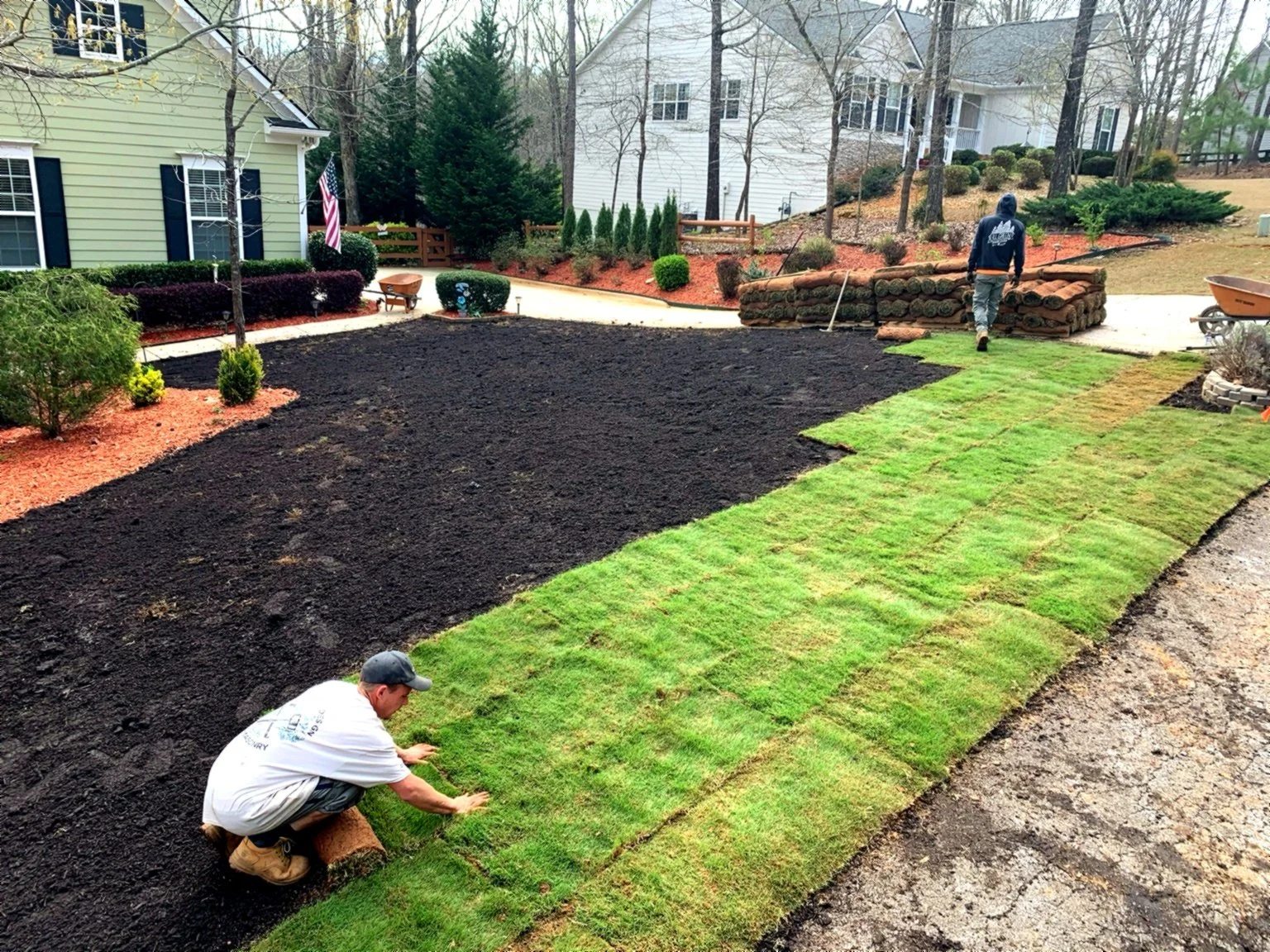 Two workers laying sod grass in a backyard yard with a house in the background.