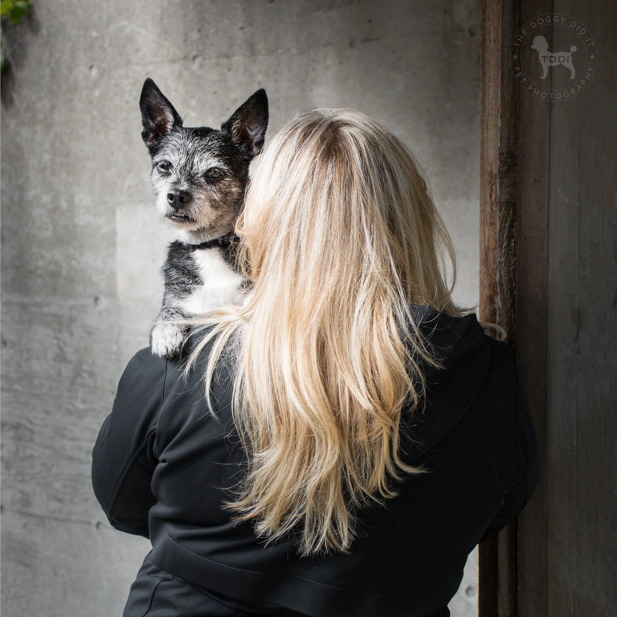 A woman with long blonde hair holding a small dog with black and white fur near her shoulder, both looking toward the camera against a gray wall.