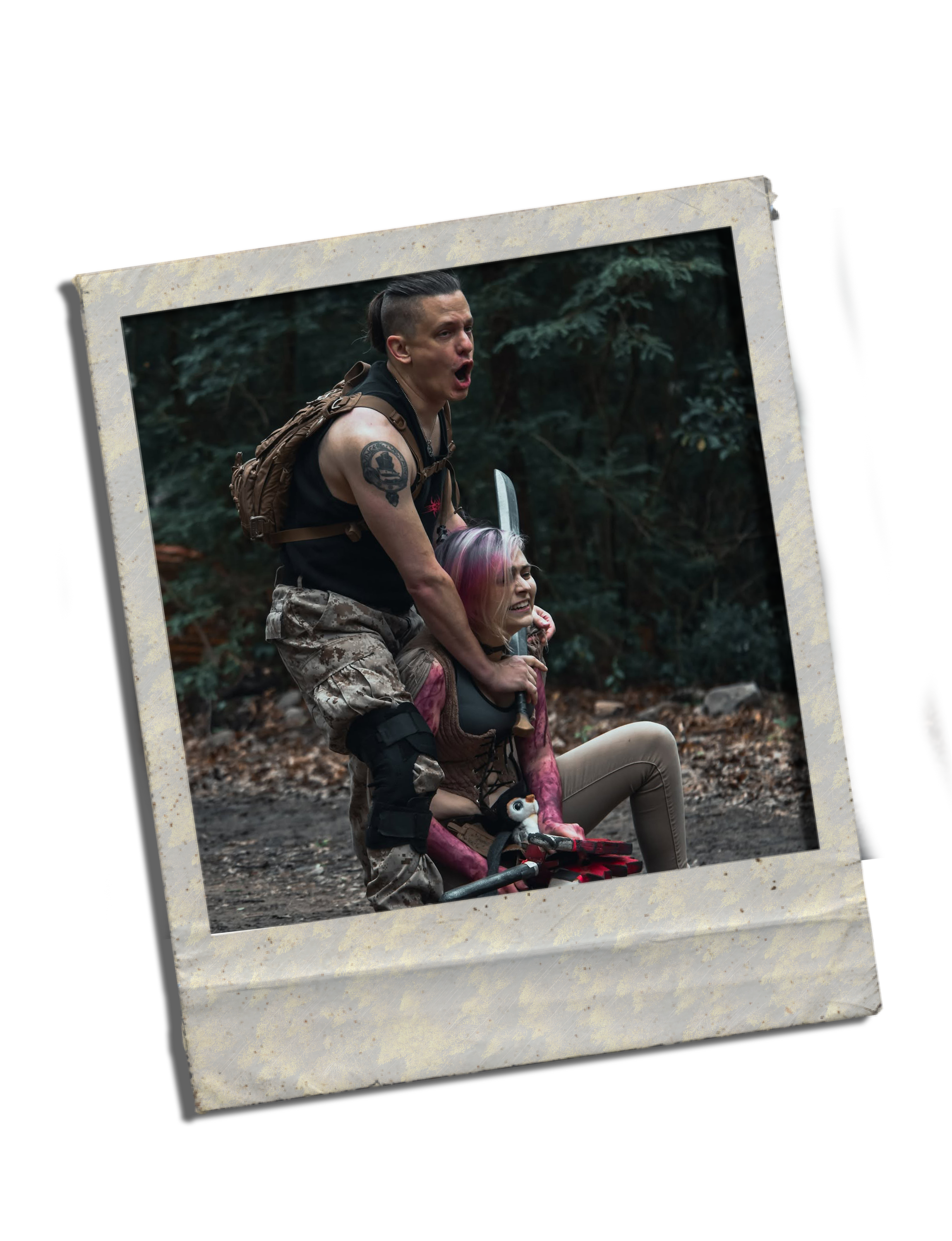 Two women in the woods, one sitting and the other standing, both with pink and purple hair, surrounded by dense greenery and trees, appearing to be engaged in a playful or animated interaction.