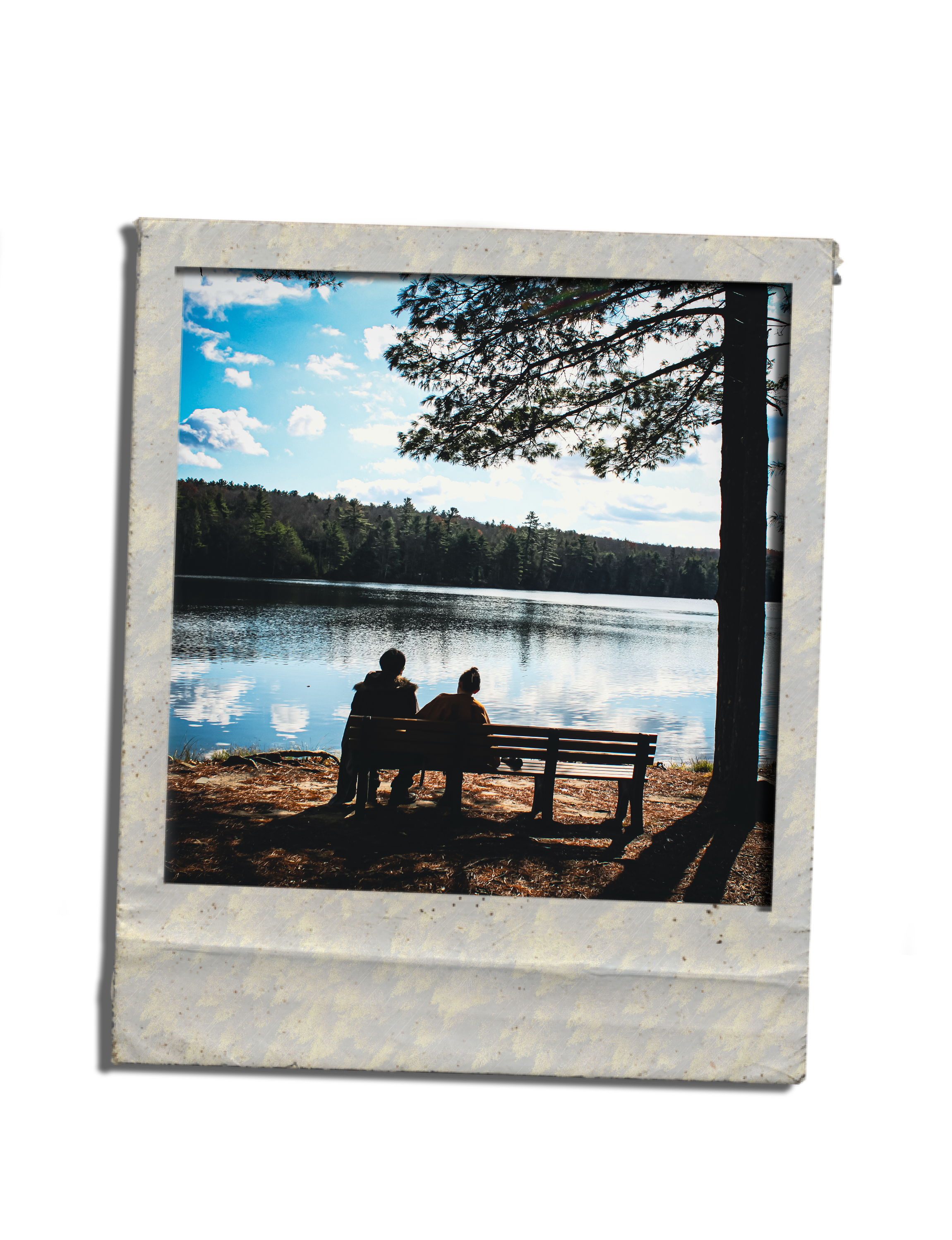Two people sitting on a park bench by the water, facing a lake with trees and a blue sky with clouds.